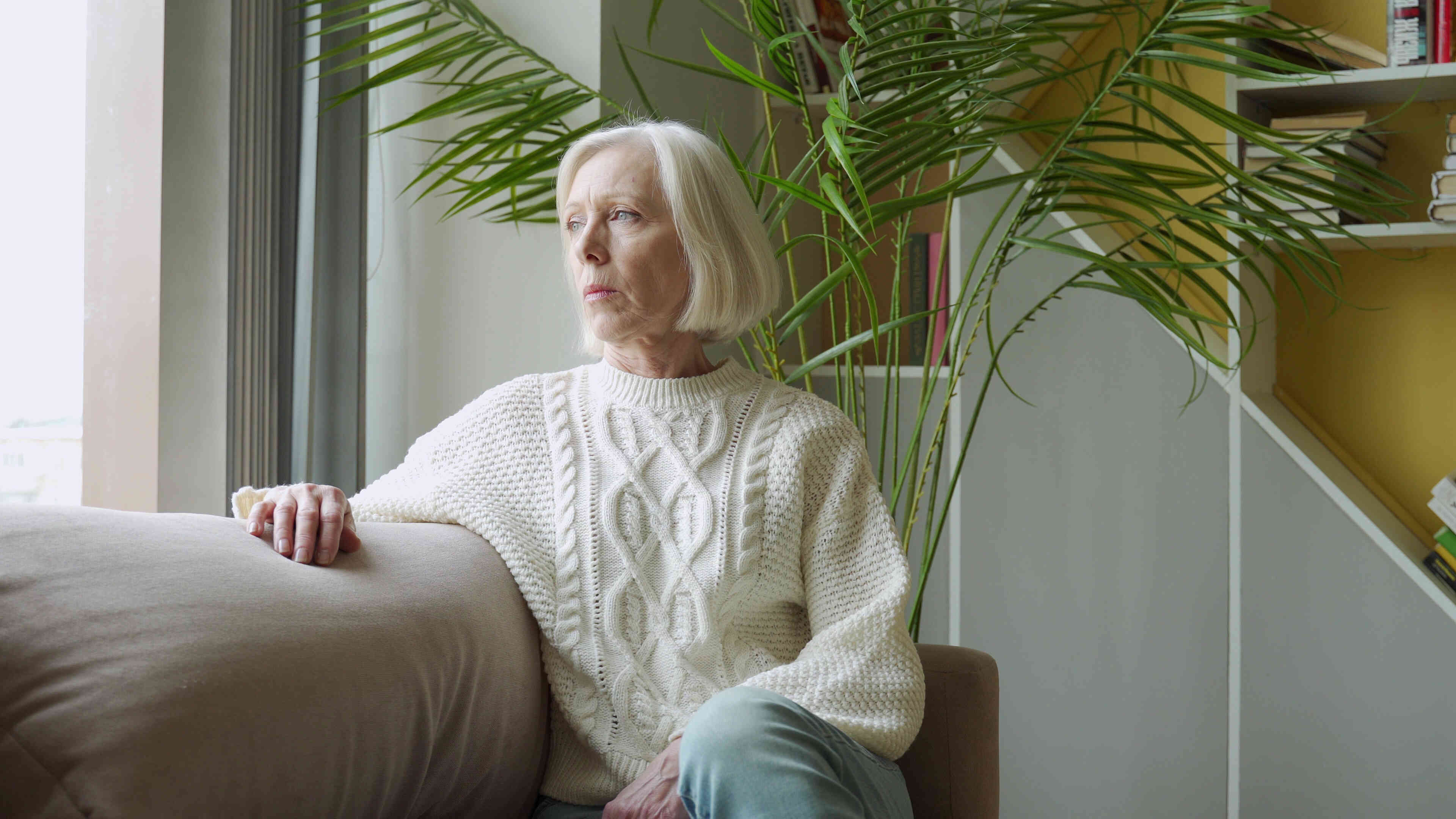 A mature woman in a white knit sweater sits on her couch and gazes off with a sad expression.