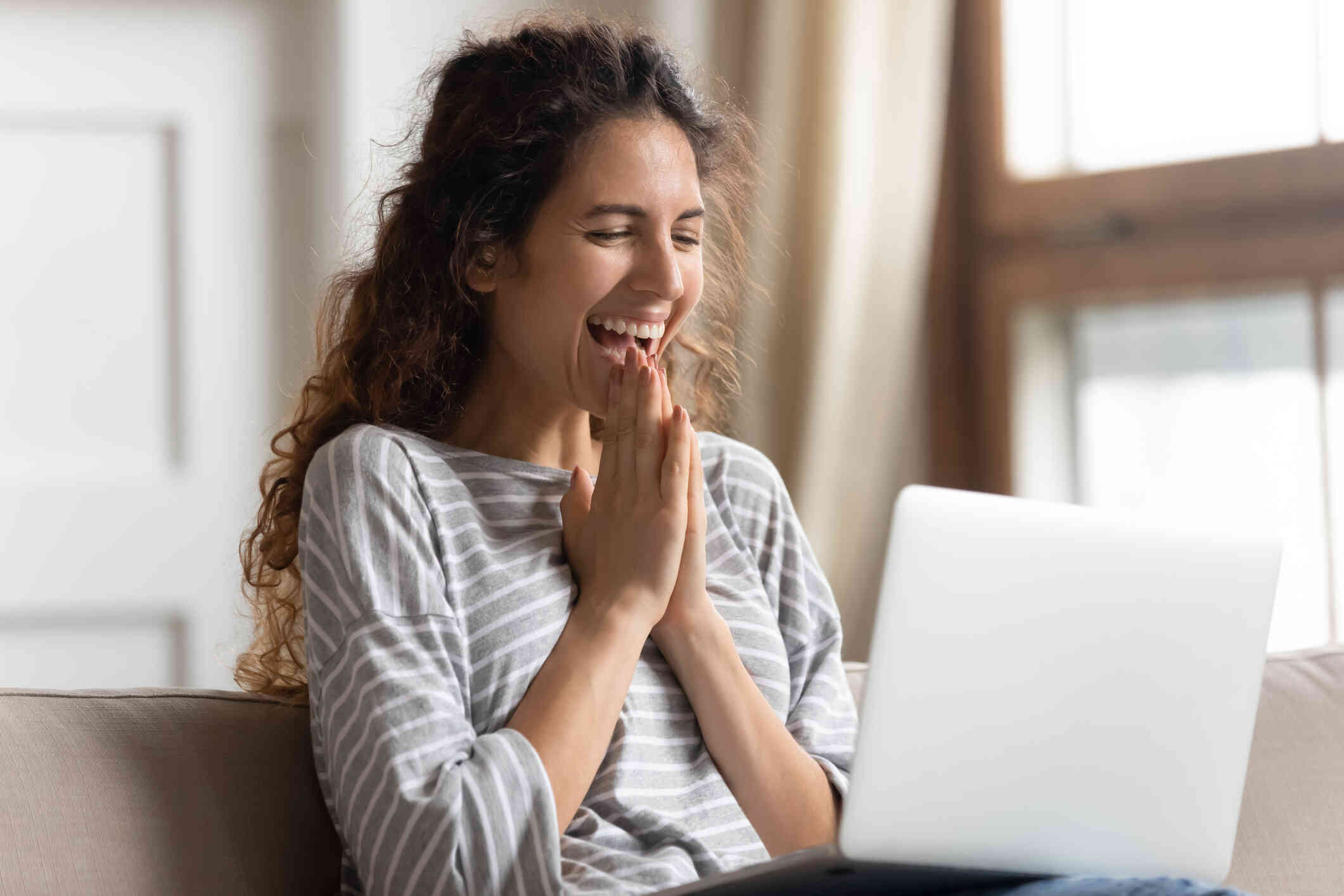 A woman sits at a table and looks at the laptop open infront of her with an excited expression.