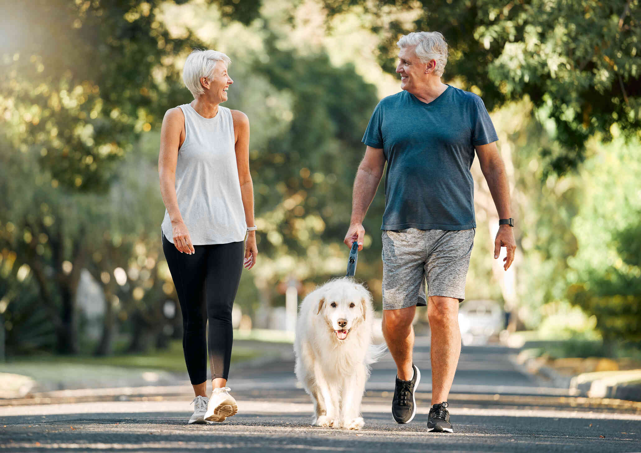 A mature man and woman wearing athletic clothes walk outside down a path with trees. The man walks a white fluffy dog who is on a leash.