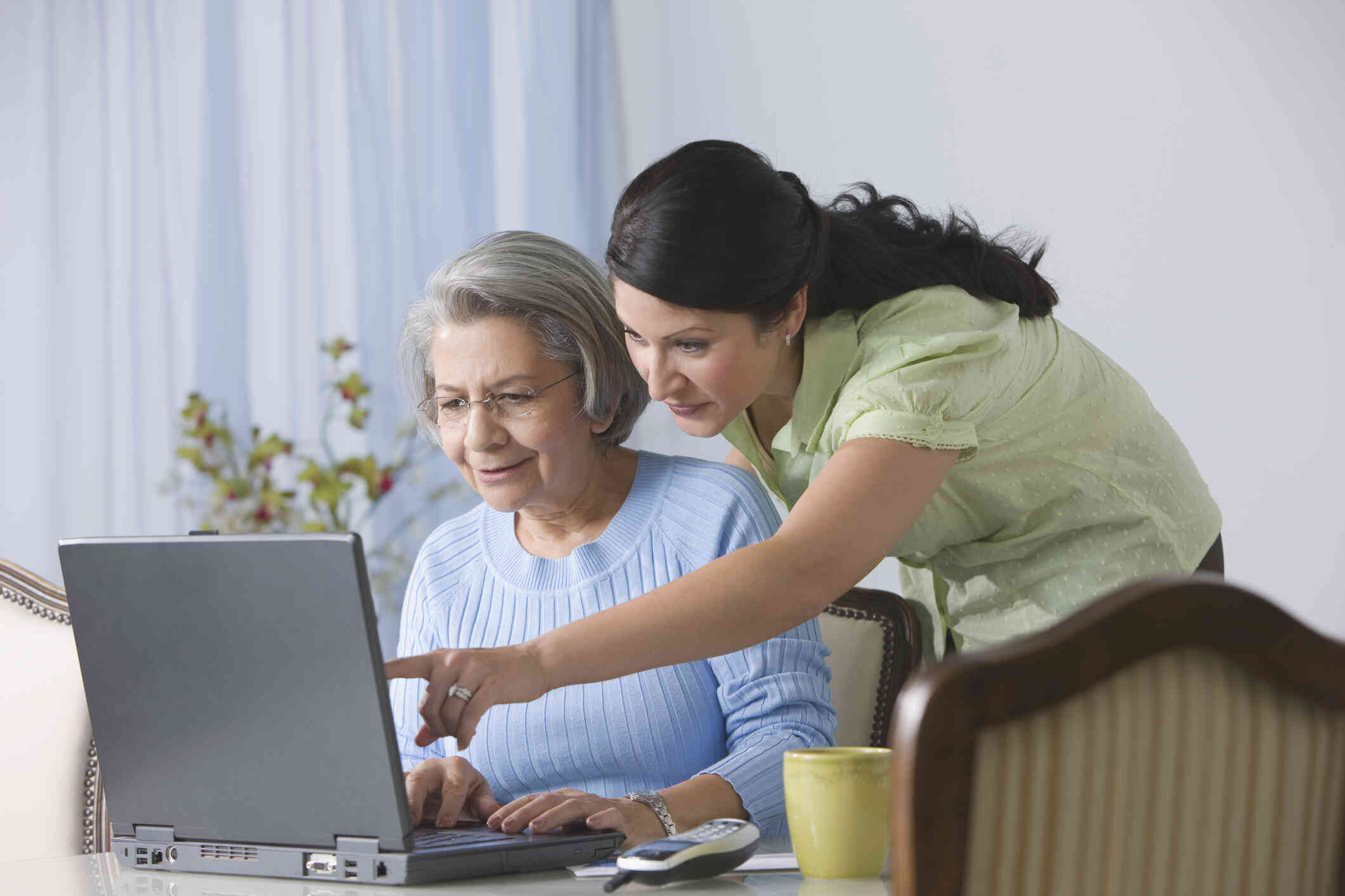 An elderly woman in a blue sweater sits at the kitchen table with a laptop open infront of her as another women standing behind her leans over to point at the laptop screen.