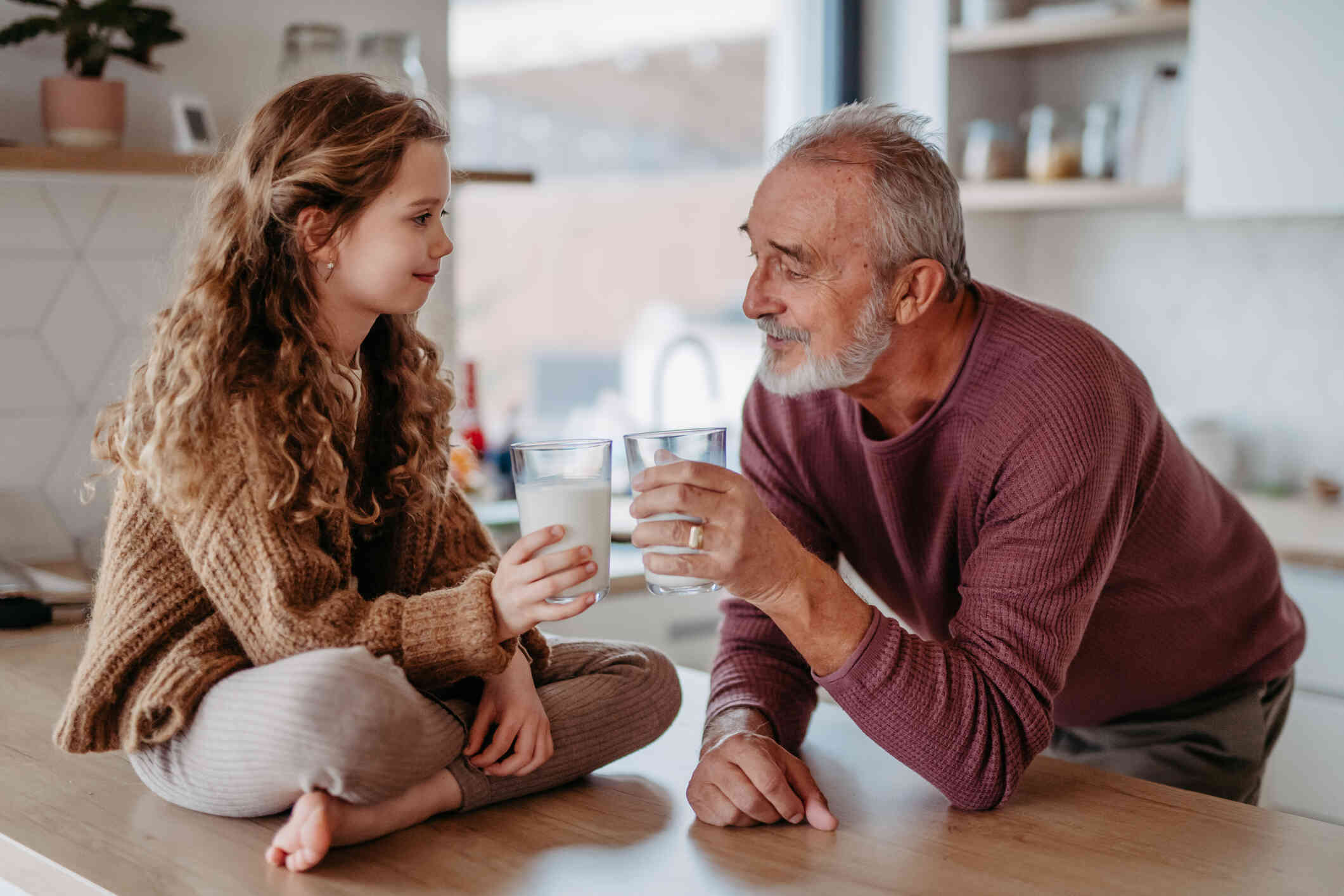 A young girl sits on a kitchen counter with a glass of milk as her dad leans on the counter with his glass of milk and cheers the glasses together.