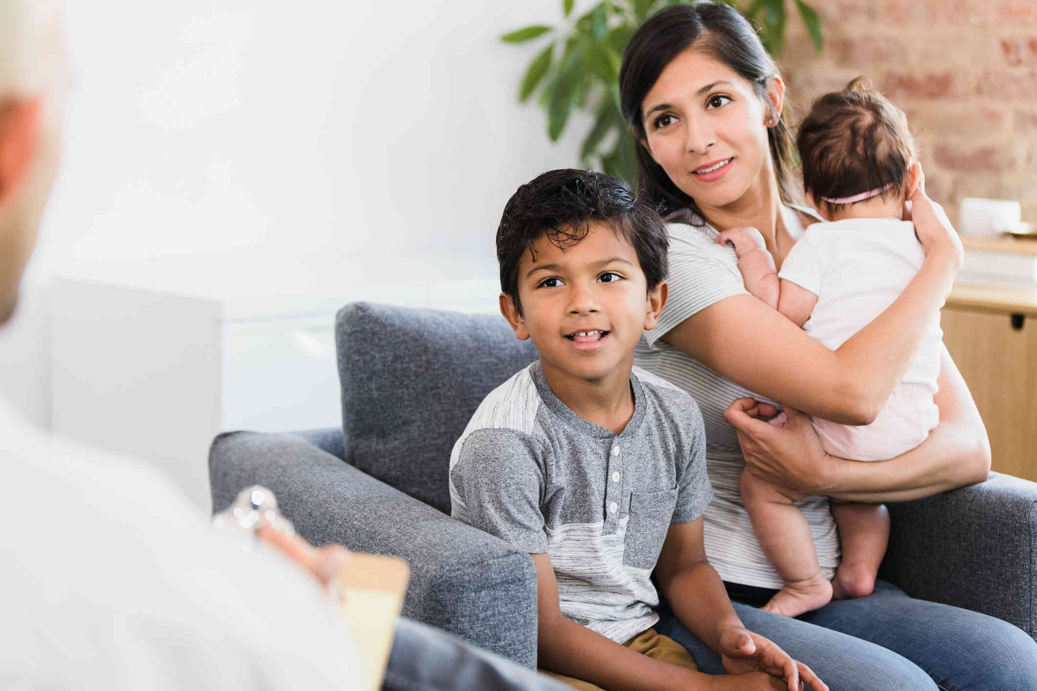 A mother holds her infant daughter in her arms while sitting in a chair with her young son as they smile at the therapist sitting across from them.