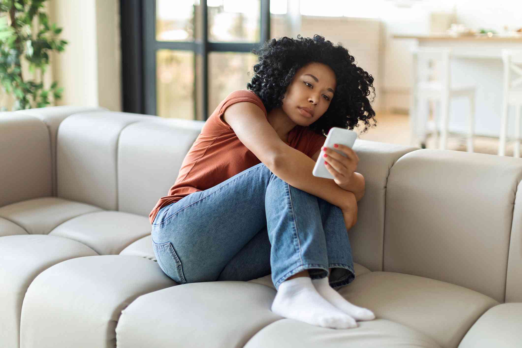 A woman in an orange shirt sits curled up on a grey couch and looks sadly down at the cellphone in her hand.