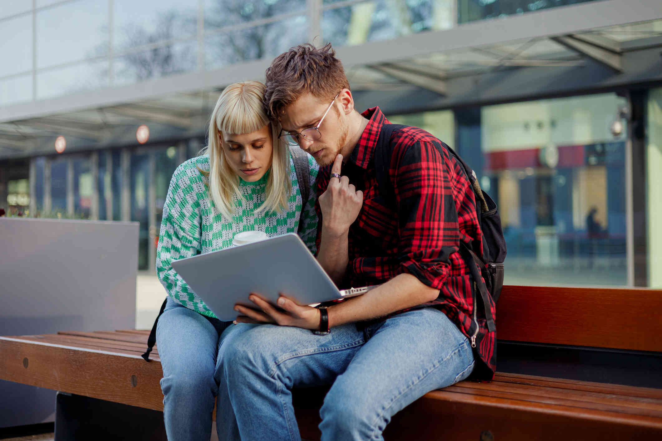 A couple seated on an outdoor bench, both focused on the laptop screen.