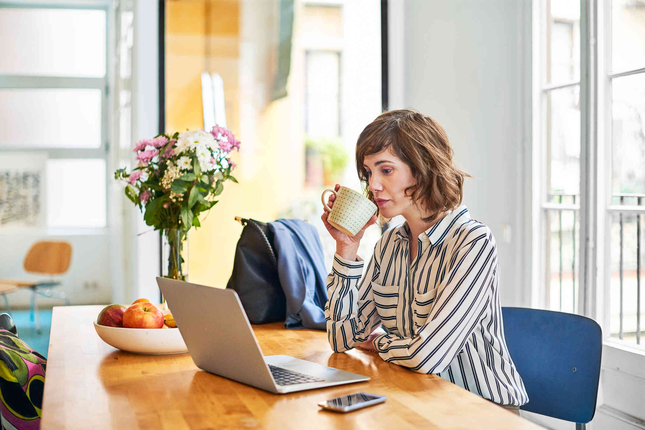 A woman in a striped shirt sits at the kitchen table and drinks from a cup of  coffee while looking at the open laptop infront of her.