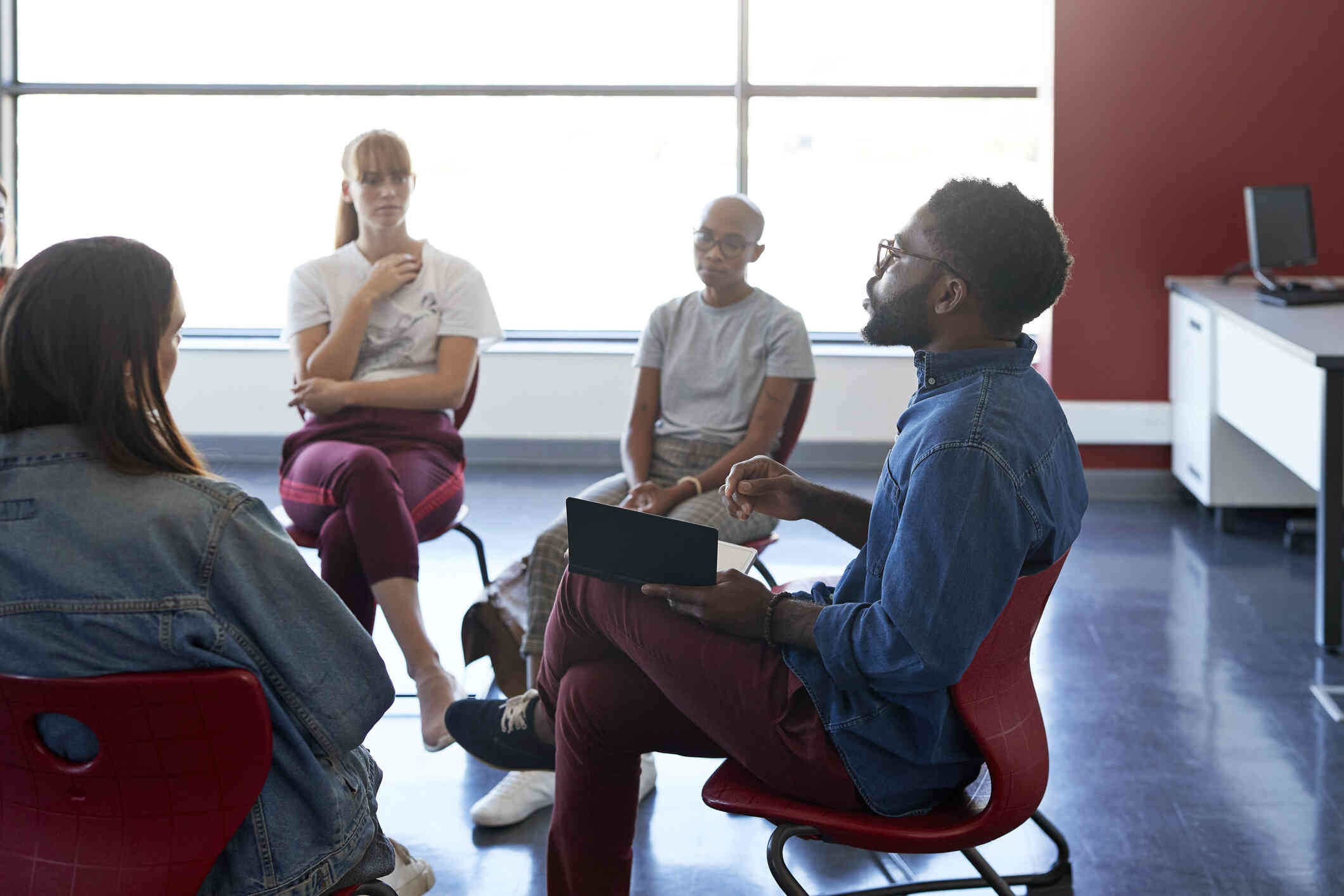 A group of adults sit in a group therapy circle led by a male therapist with a clipboard. 