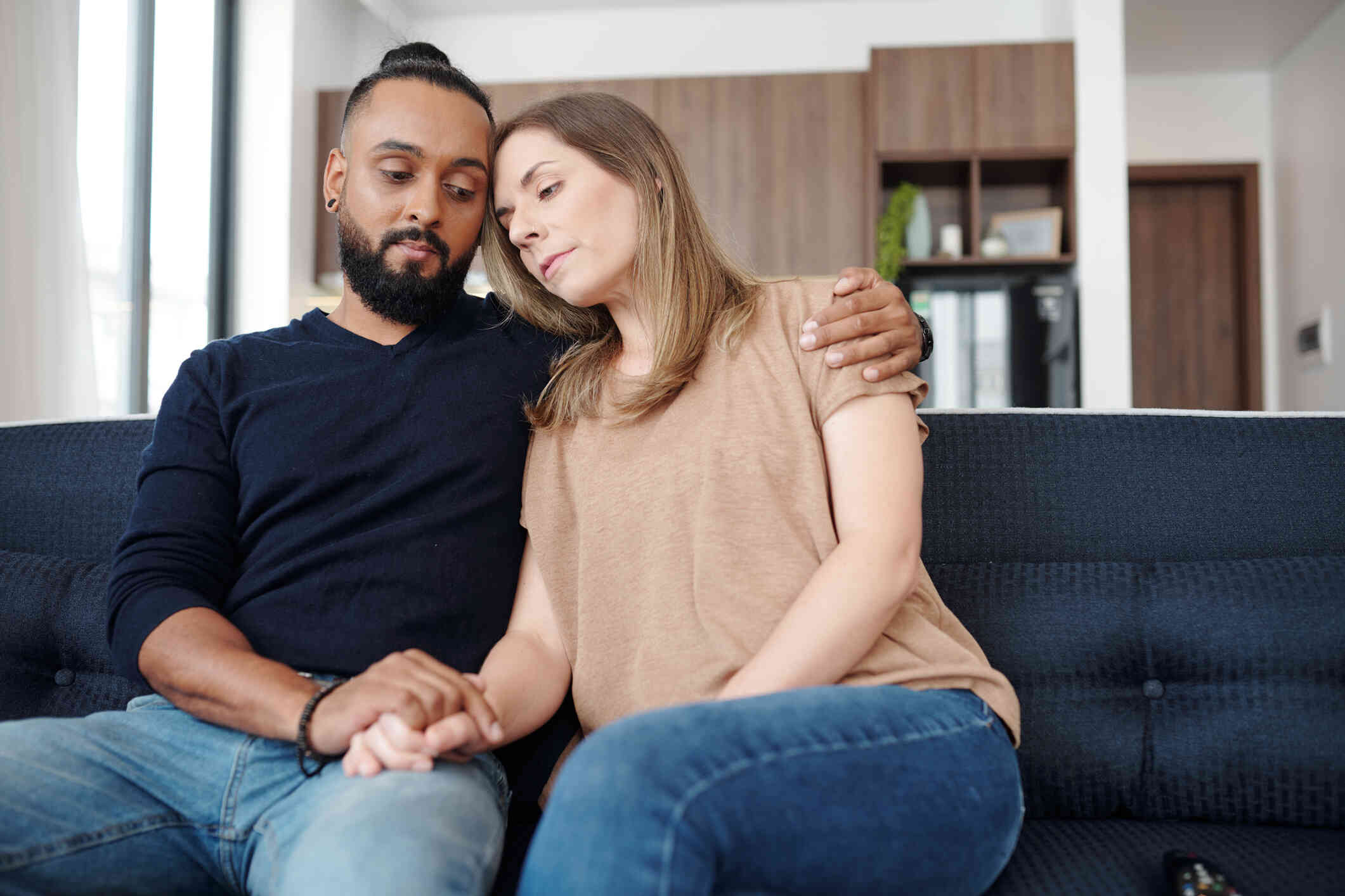 A man sits on the couch and wraps his arm around his female partner sitting next to him with a serious expression.