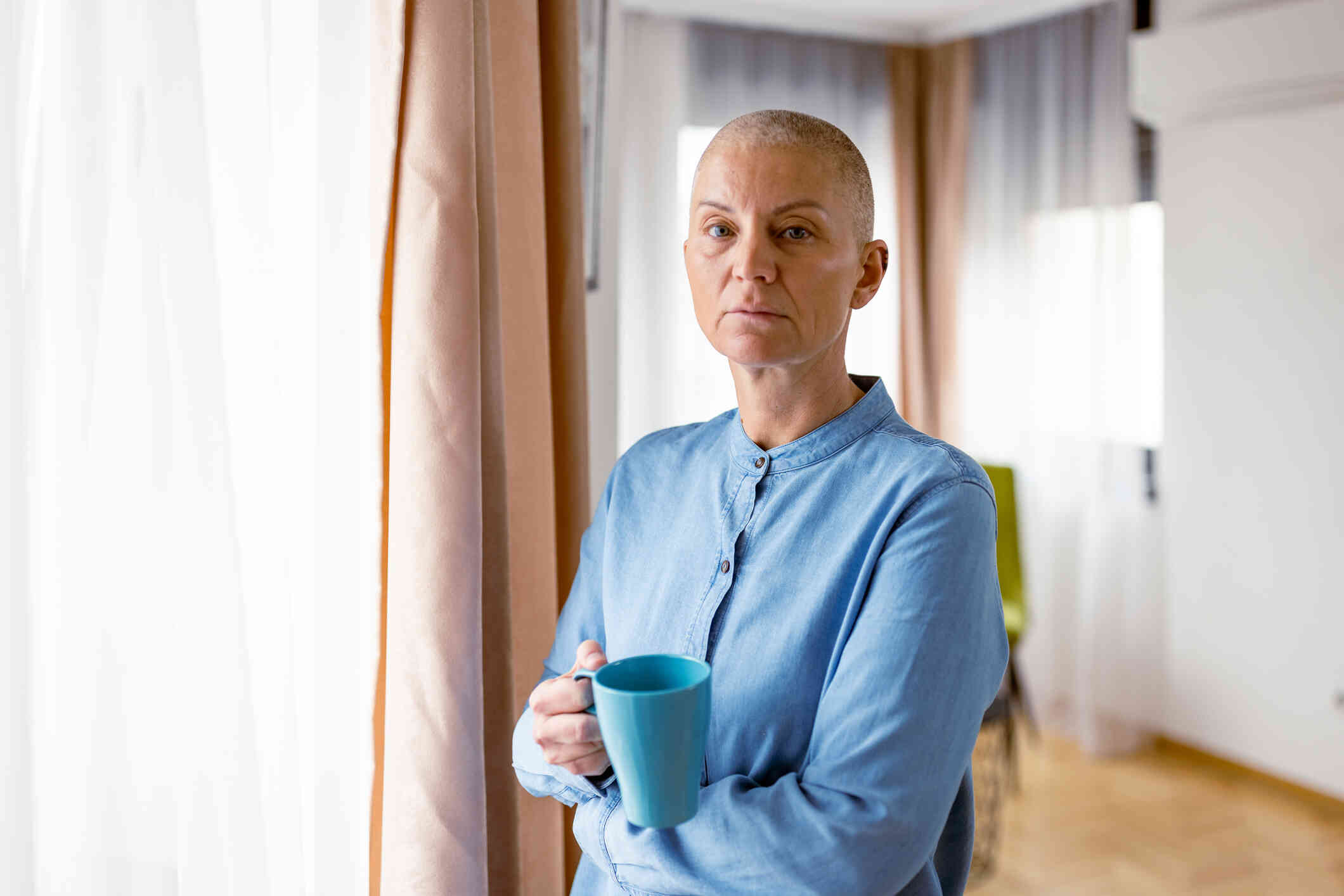 A woman in a blue shirt stands in her home while holding a coffee mug and loks at the camer awith a serious expression.