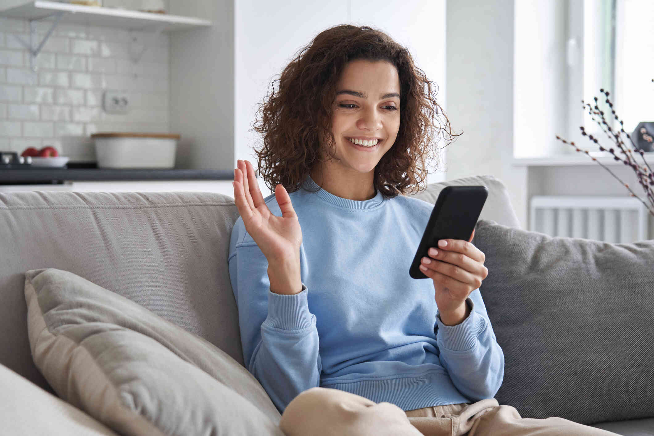 A girl in a blue sweater sits on the couch and waves while smiling at at the cellphone in her hand during a video call.