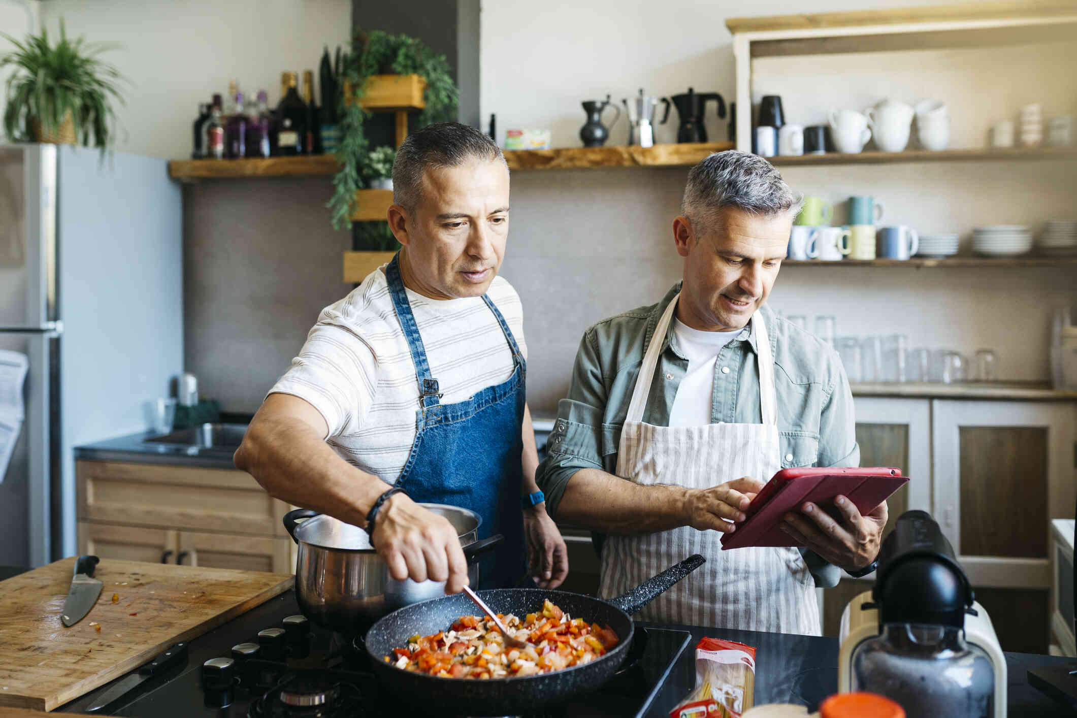 Two mature men in aprons stand over a stovetop cooking inside a home.