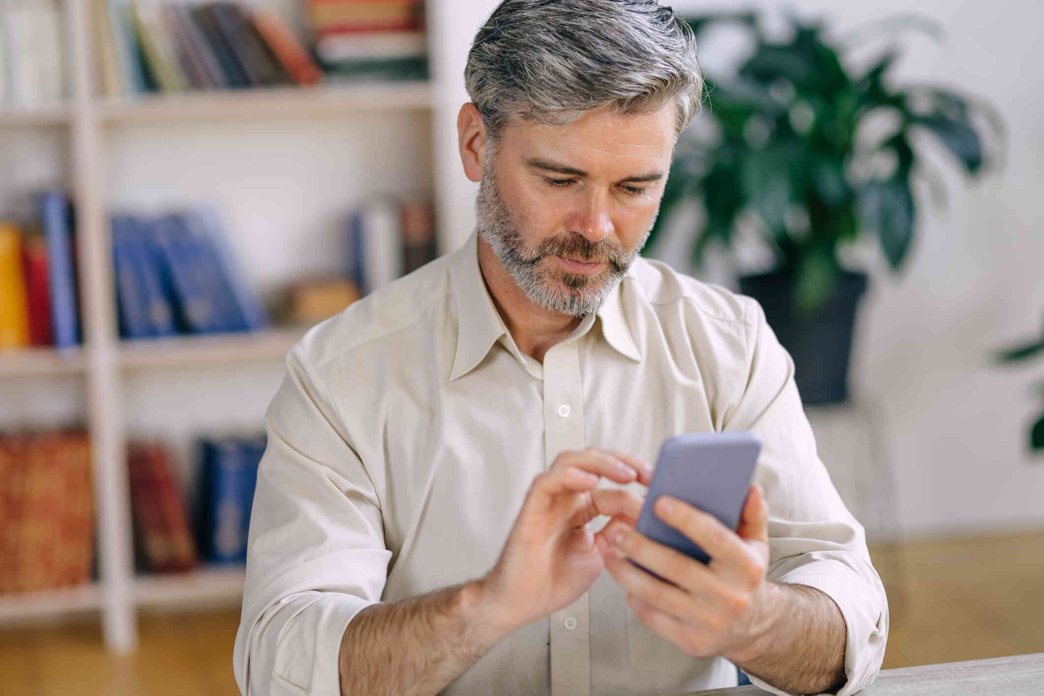 A middle aged man in a button down shirt sits head a bookcase and looks down at the cellphone in his hand.