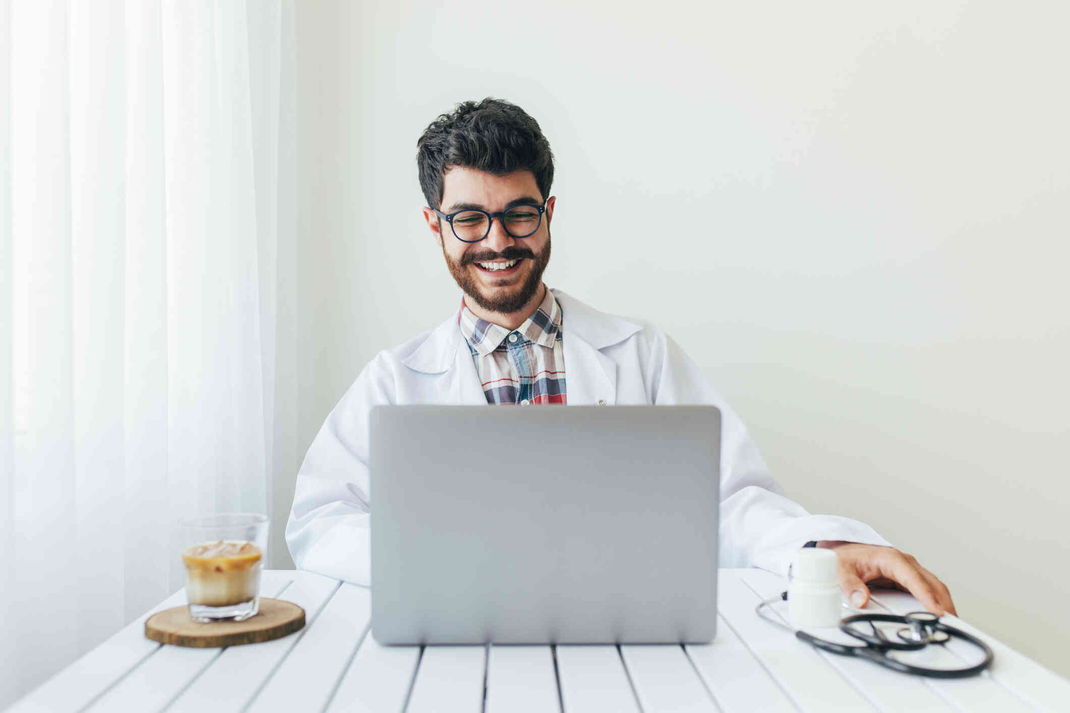 A young therapist smiles while looking at his laptop screen, seated at a table.