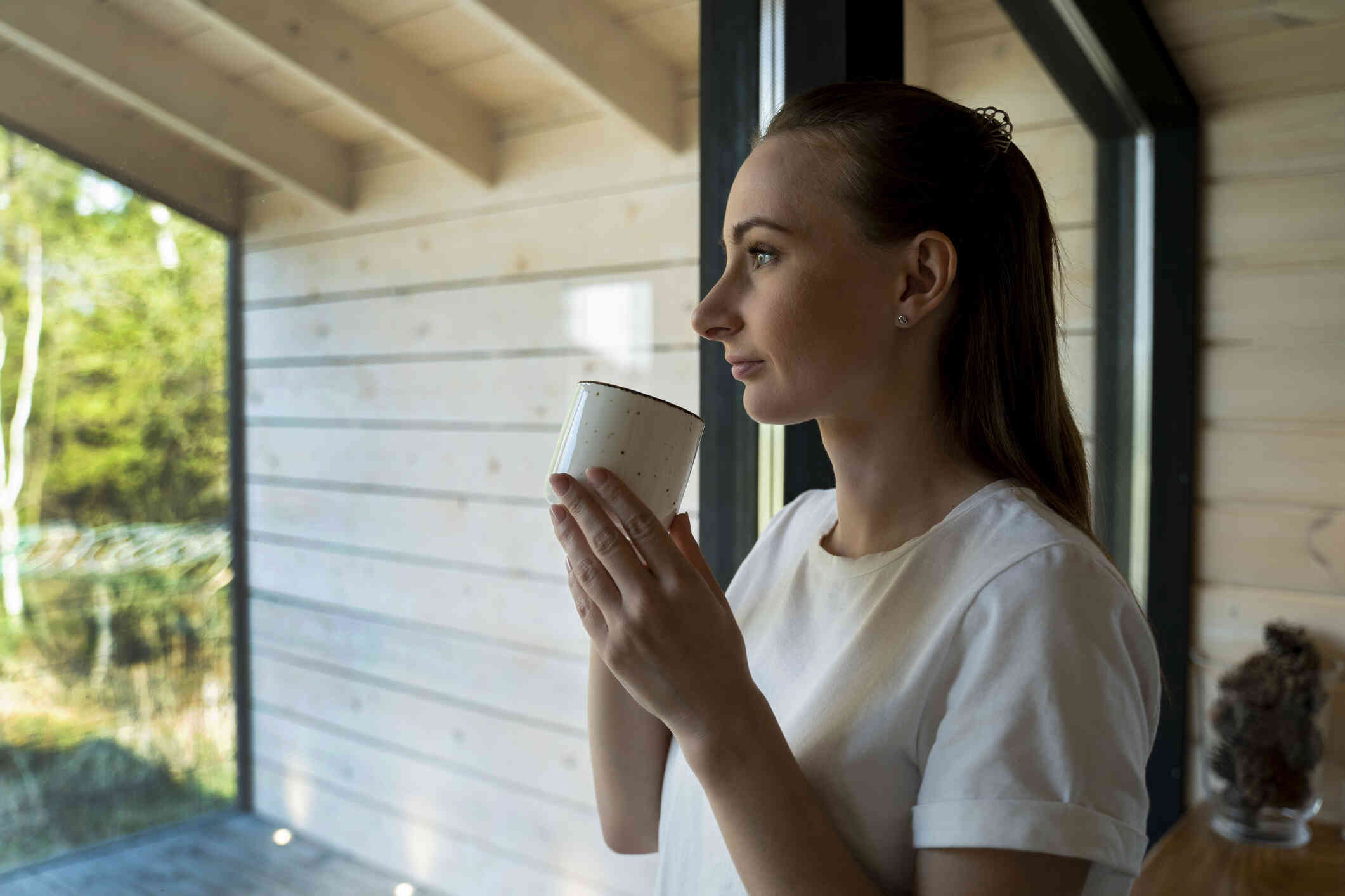 A woman in a white shirt stands near a window in her home and holds a white coffee mug near her face as she gazes out of the window while deep in thought.