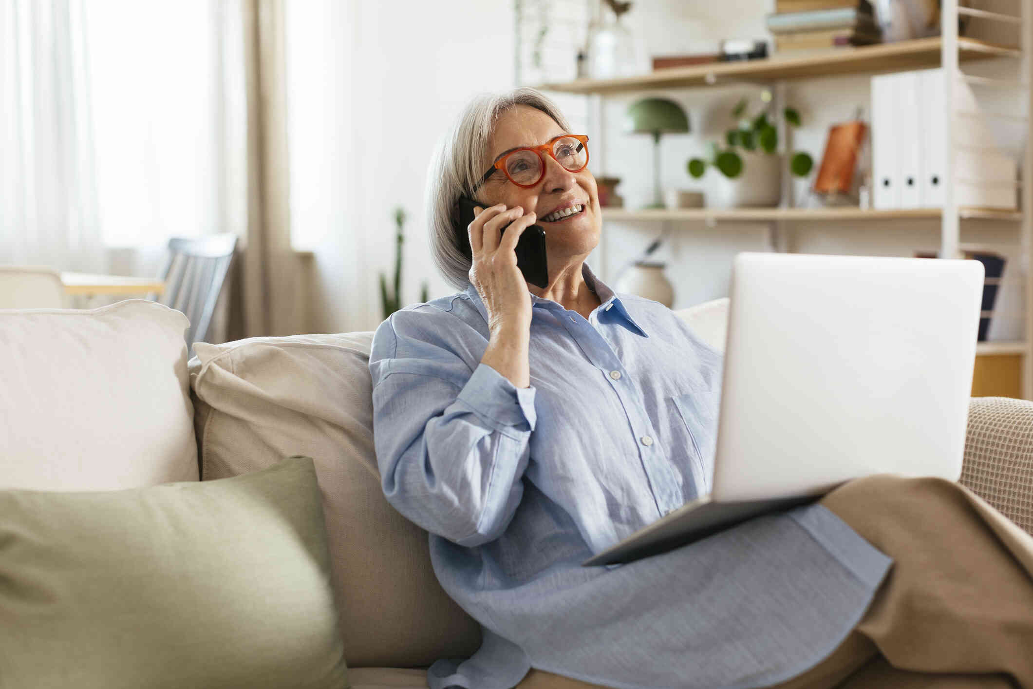 A mature woman with red glasses sits on the couch with her laptop in her lap and talks on her cellphone while smiling. 