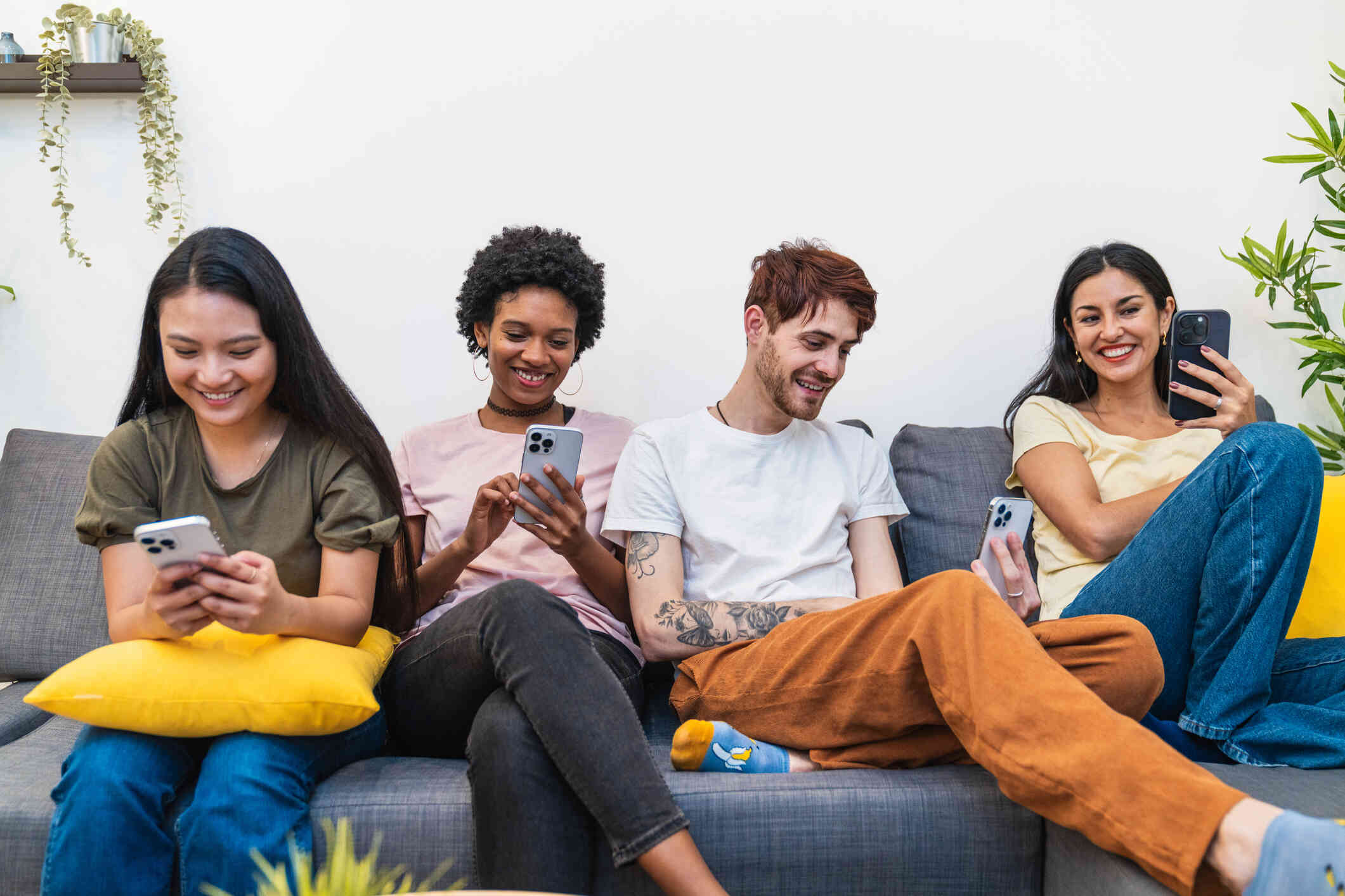 Three young women and one man with tatoos on his arm sit on a couch next to each other. They each hold a cell phone and look down at the screens with a smile.