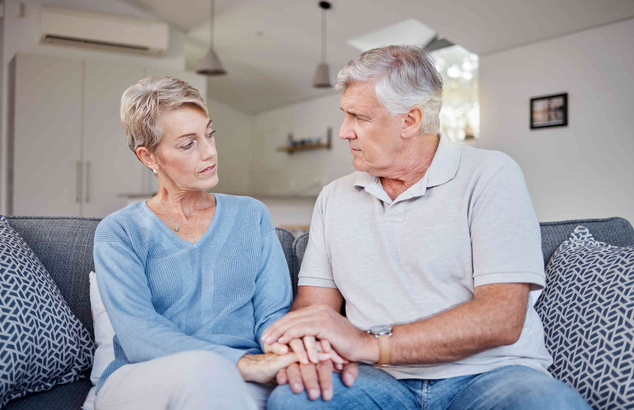 A close-up image of an elderly couple sitting on their couch, holding hands and gazing at each other lovingly.