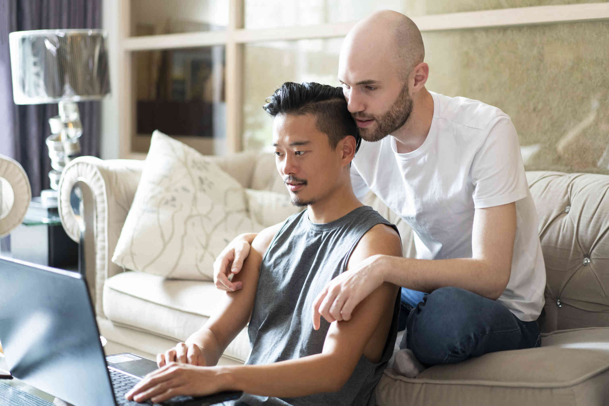 A man in a white shirt sits on a couch and puts his arms around another man in a gray tank top as the two look down at a laptop in front of them with serious expressions.