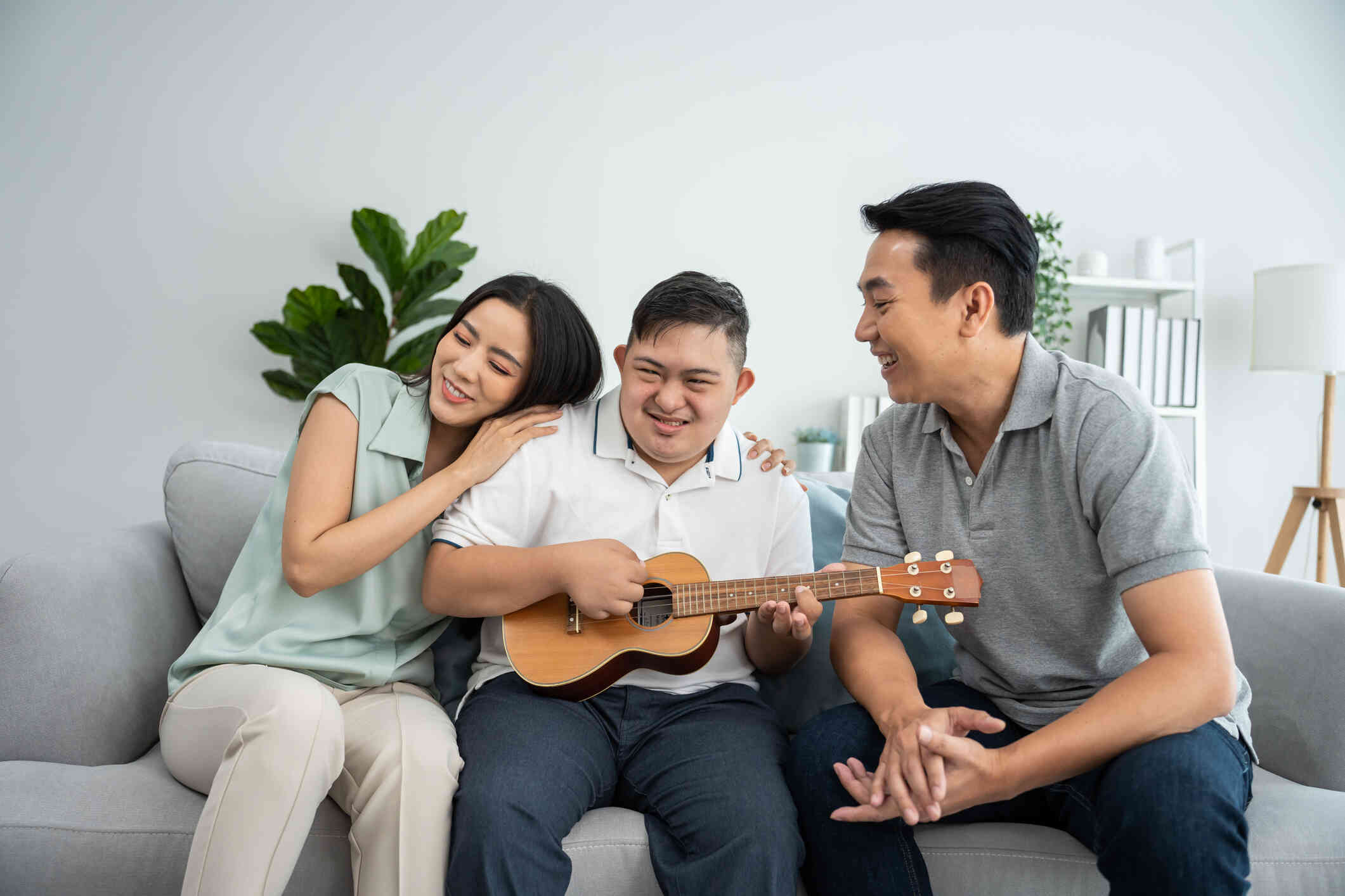 A teen boy holds a banjo while smiling as his mother and father sit happily on either side of him.