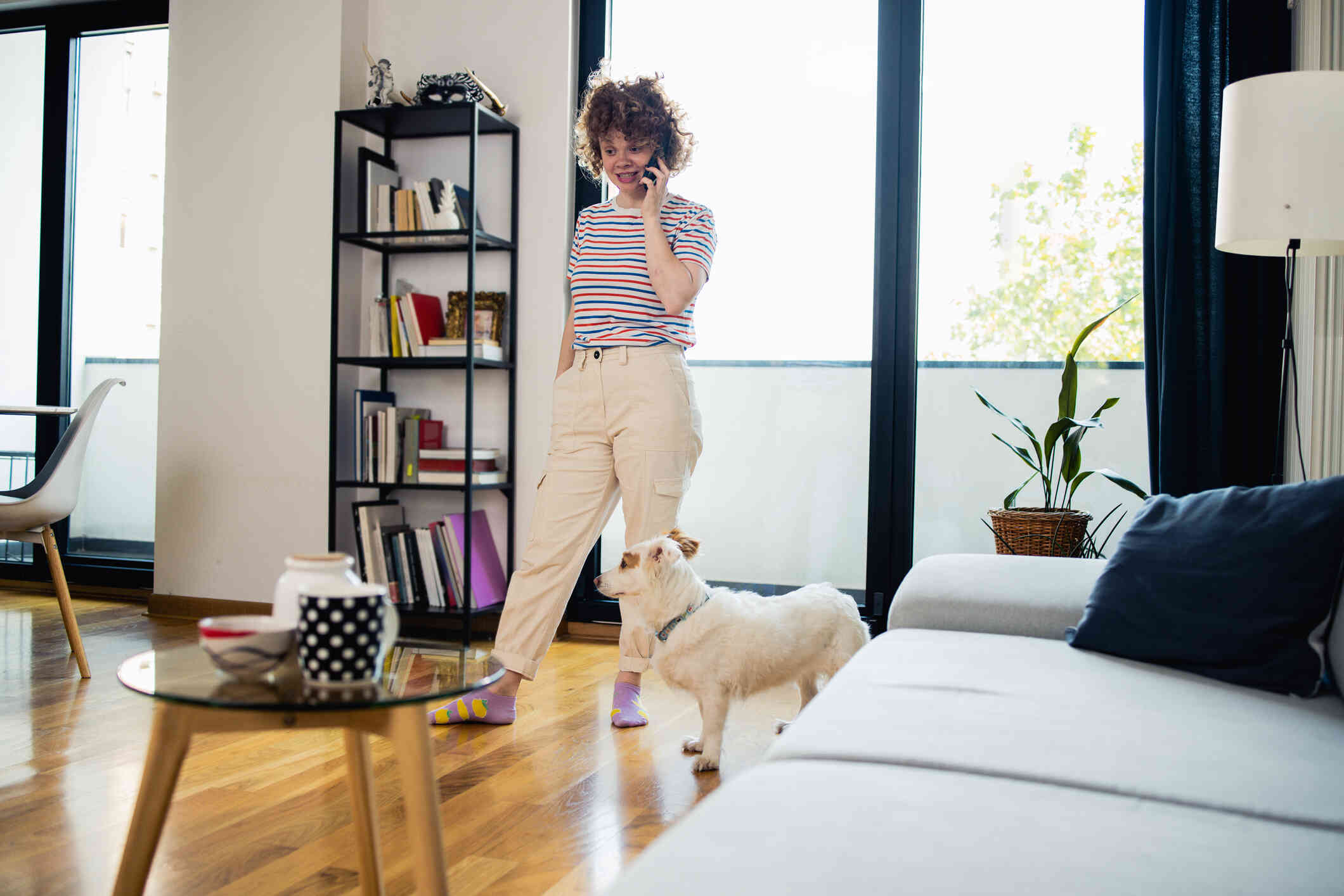 A woman in a striped shirt talks on the phone while walking around her living room with her small dog.