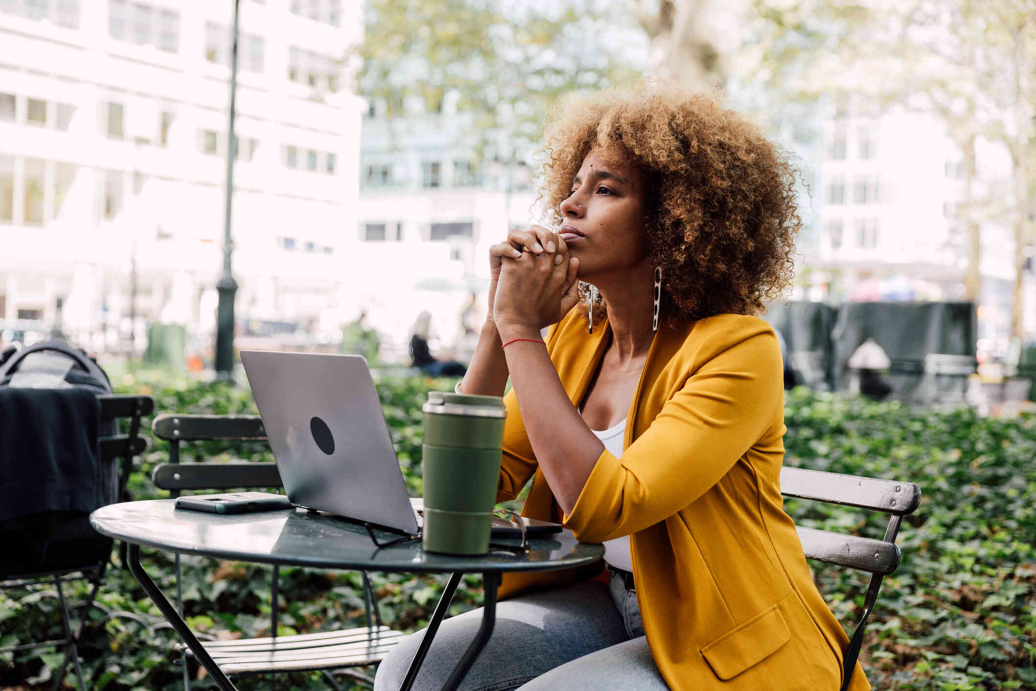 A woman in a yellow blazer sits outside at a table with her laptop and gazes off while deep in thought.