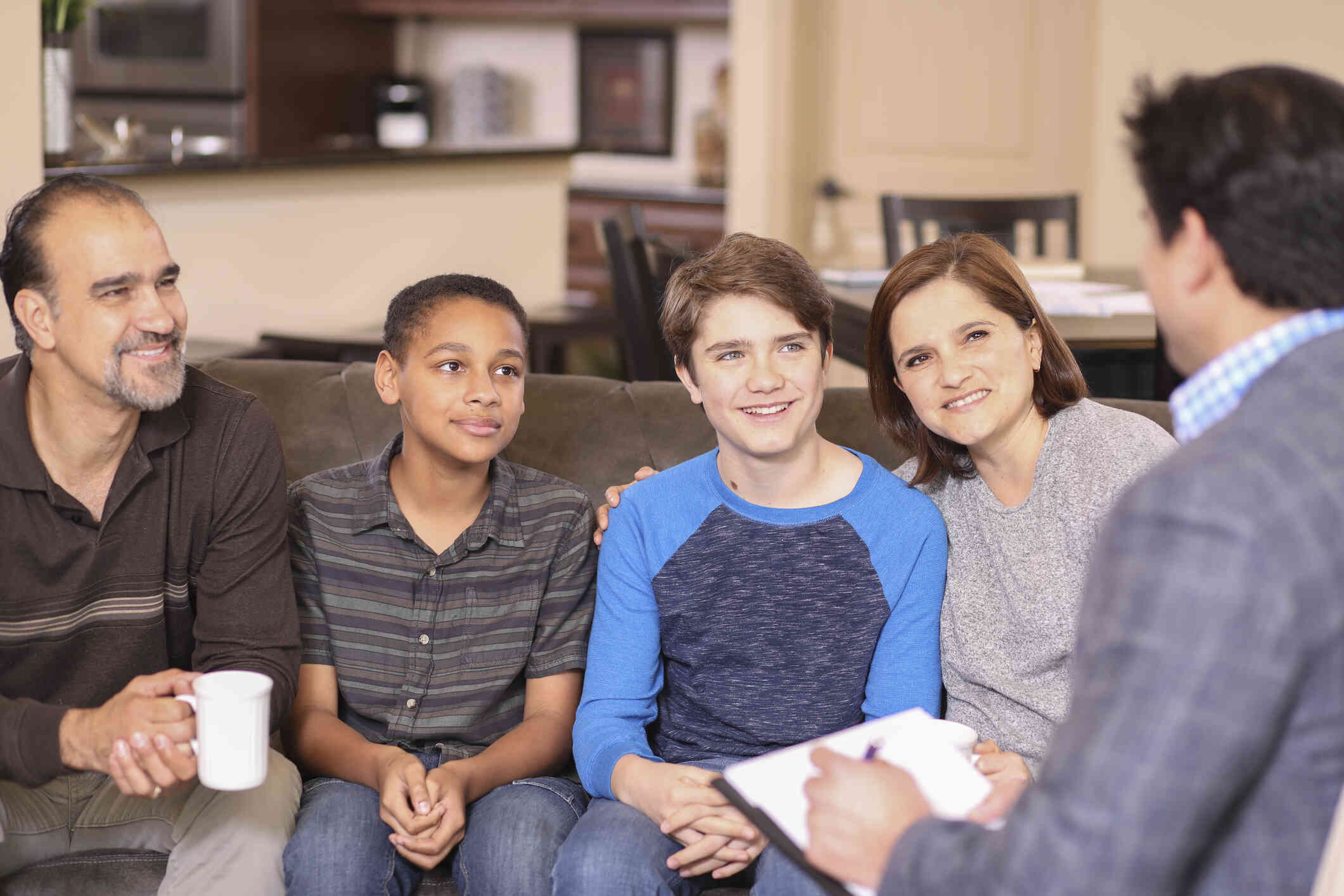 A mother and father sit on opposite sides of their two teen sons on the couch across from their family therapist during a therapy session.