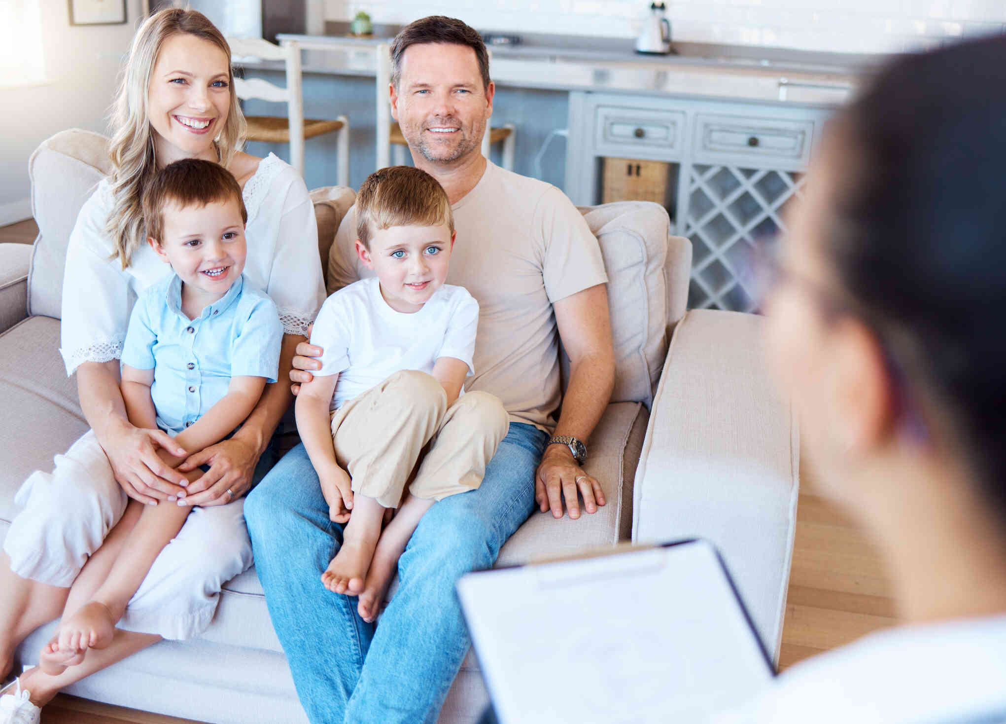 A mother and father sit on a couch with their two young sons sitting on their laps as they listen to the therapist sitting across from them during a family therapy session.