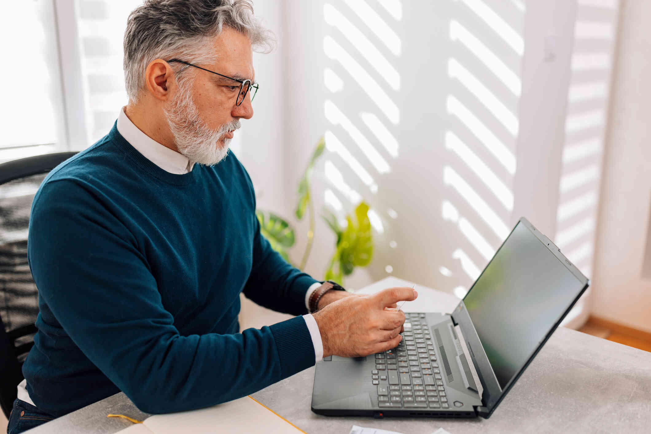 A middle aged man with glasses sits at his comptuer desk and loks at his laptop screen with a serious expression.