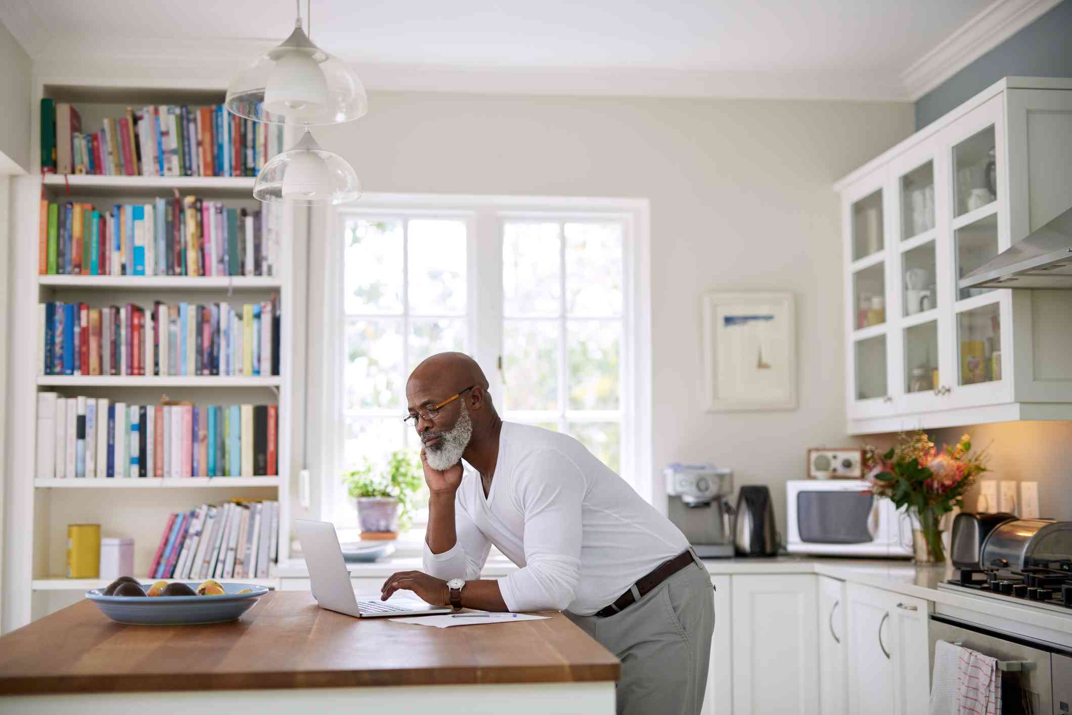A male mental health professional leans over his kitchen counter to look closely at the laptop that is open infront of him with a serious expression.