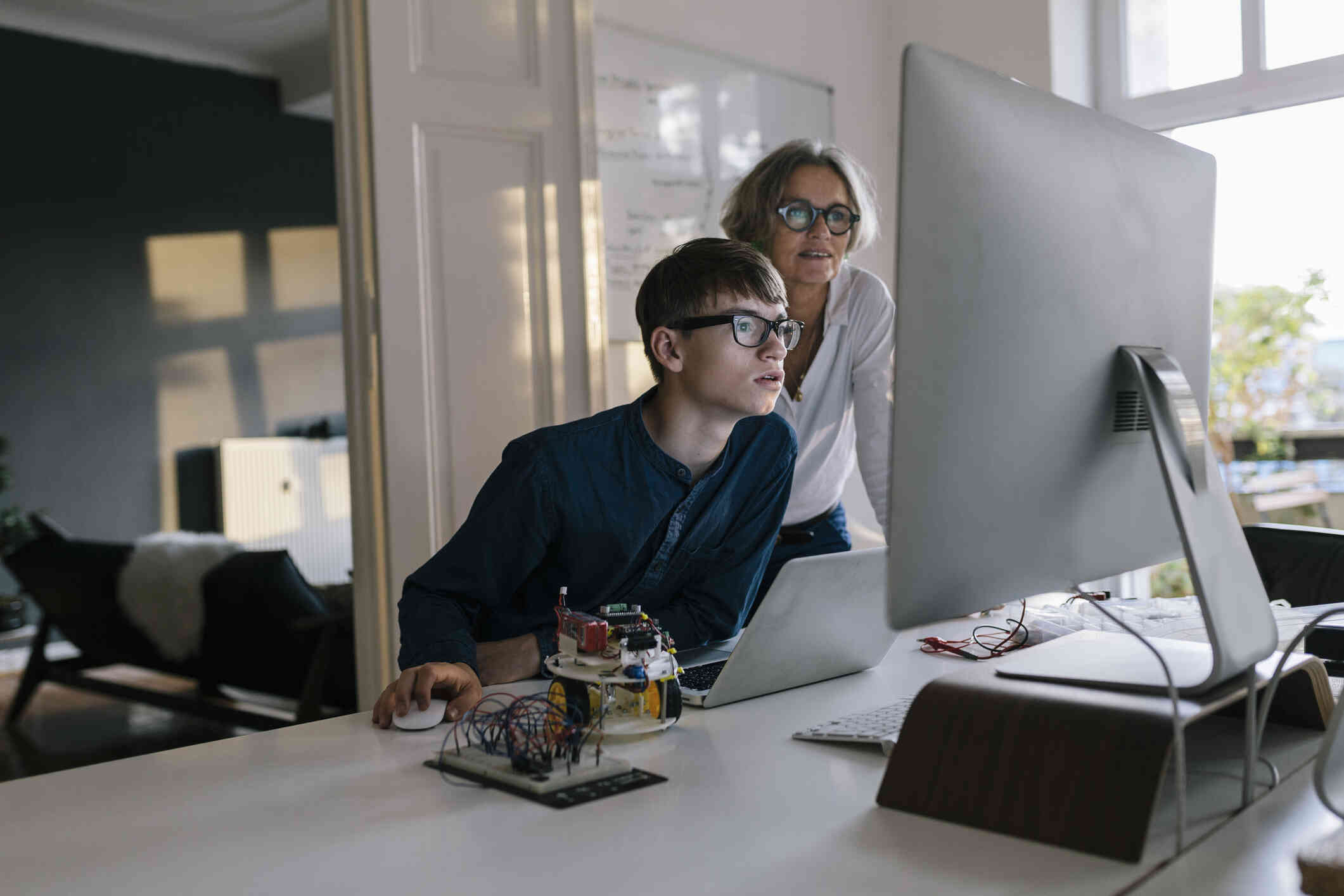 A teenage boy with glasses and his mother look intently at a desktop computer screen in their home.