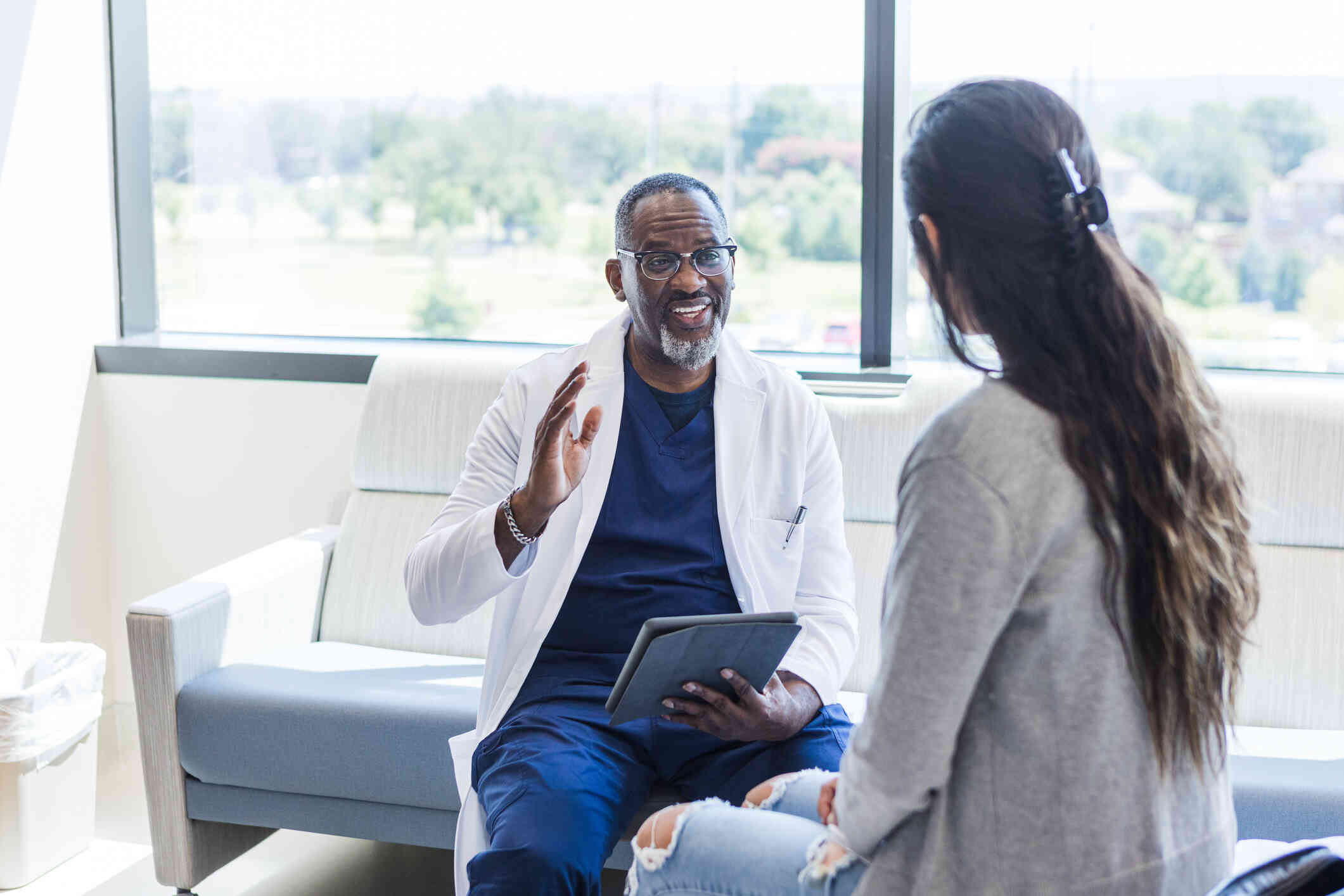 A male therapist in a white coat sits across from from his female patient and smiles while talking to her during a therapy session.