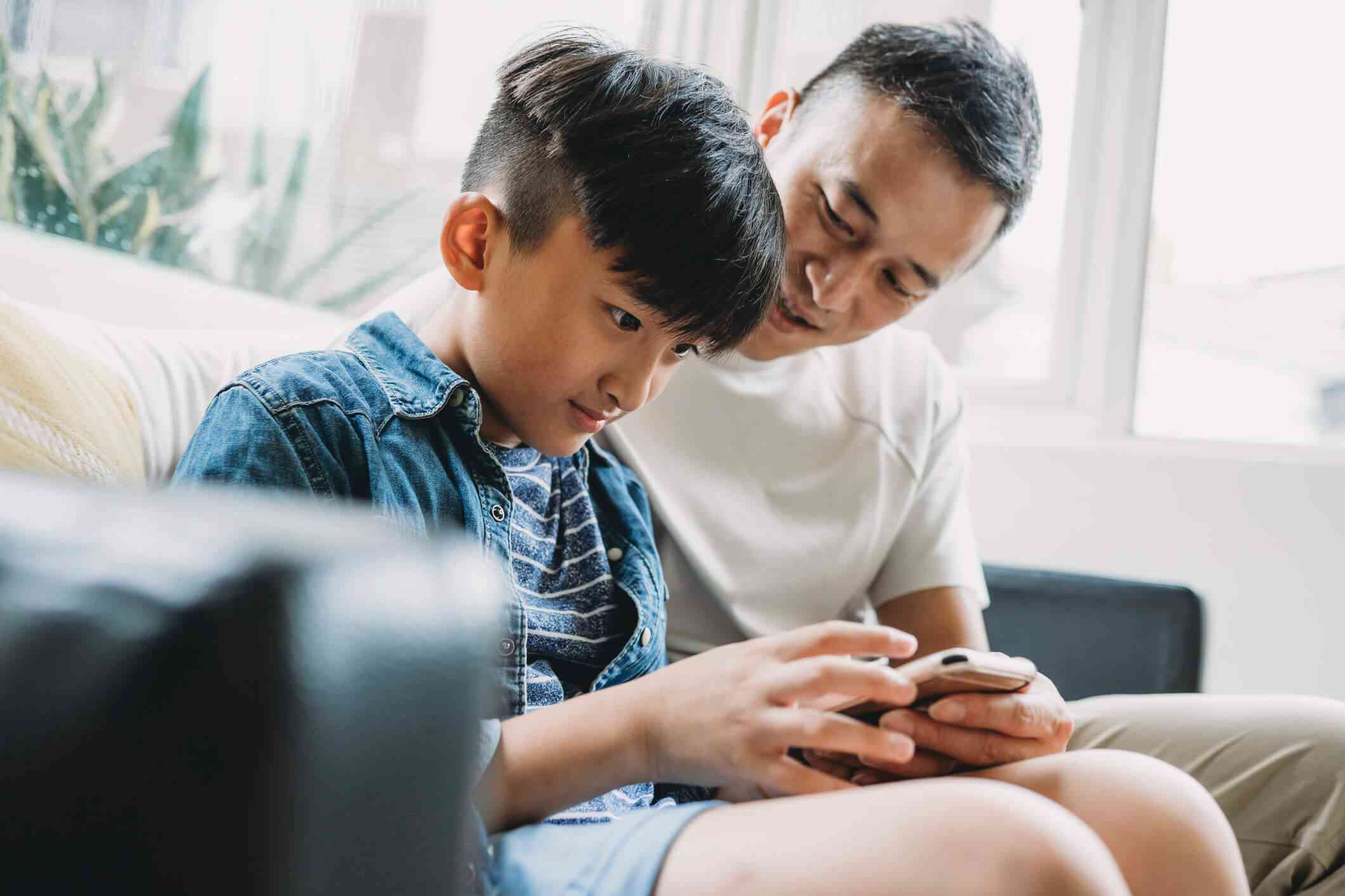 A father and his pre-teen son sit on a couch working together on a cell phone the son is holding; both are smiling. The boy is wearing blue shorts and a blue-and-white striped t-shirt under a jean jacket, and the father is wearing a white t-shirt and tan pants.