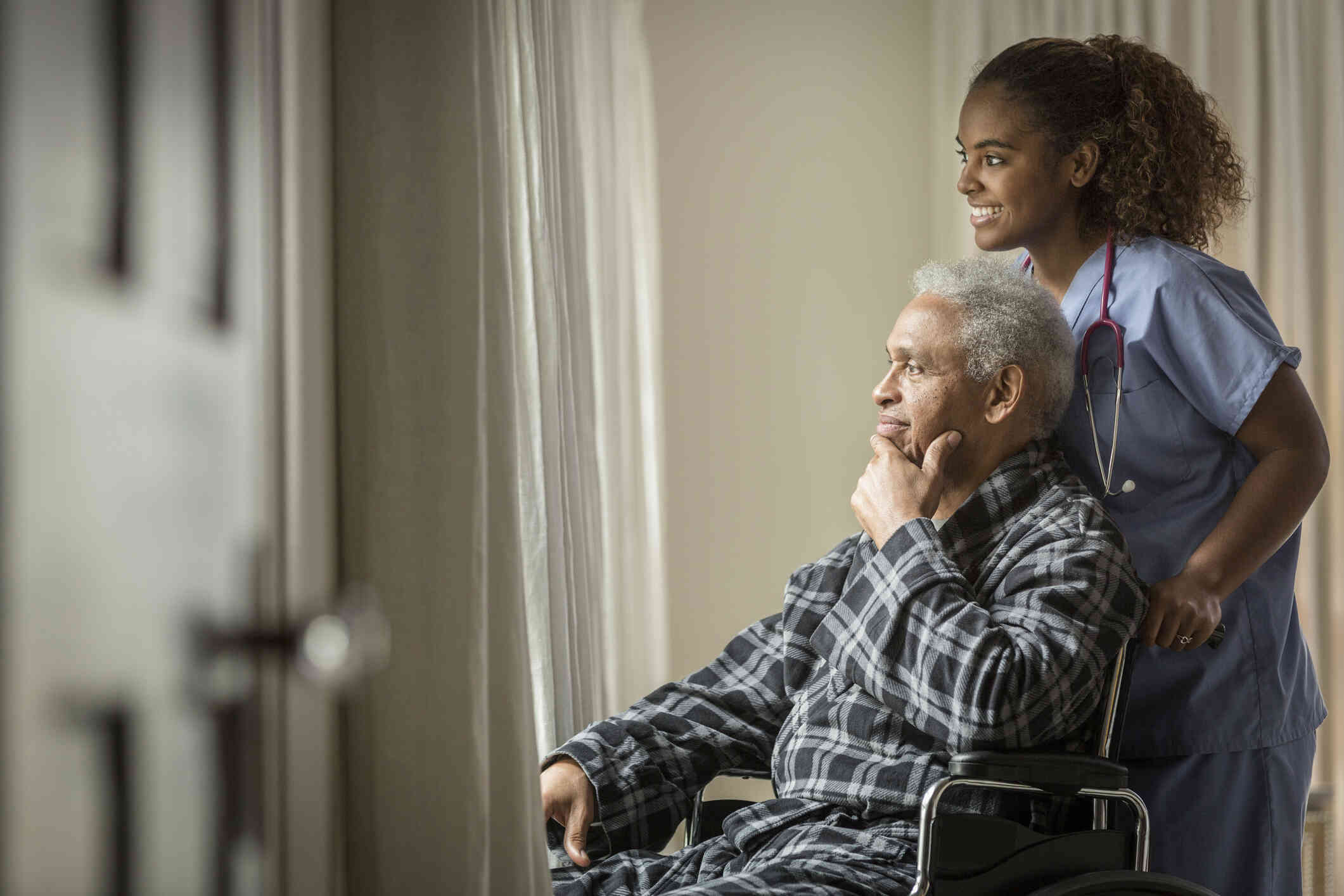 A man in wheelchair is assisted by a nurse, and they both look outside through the glass.