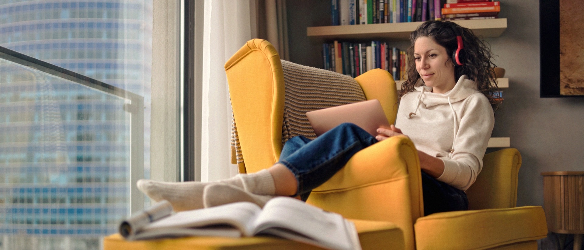 Mujer joven con auriculares usa un portátil relajada en un sillón amarillo junto a la ventana. El apoyo psicológico online puede ayudarte a gestionar la ansiedad social a tu ritmo.