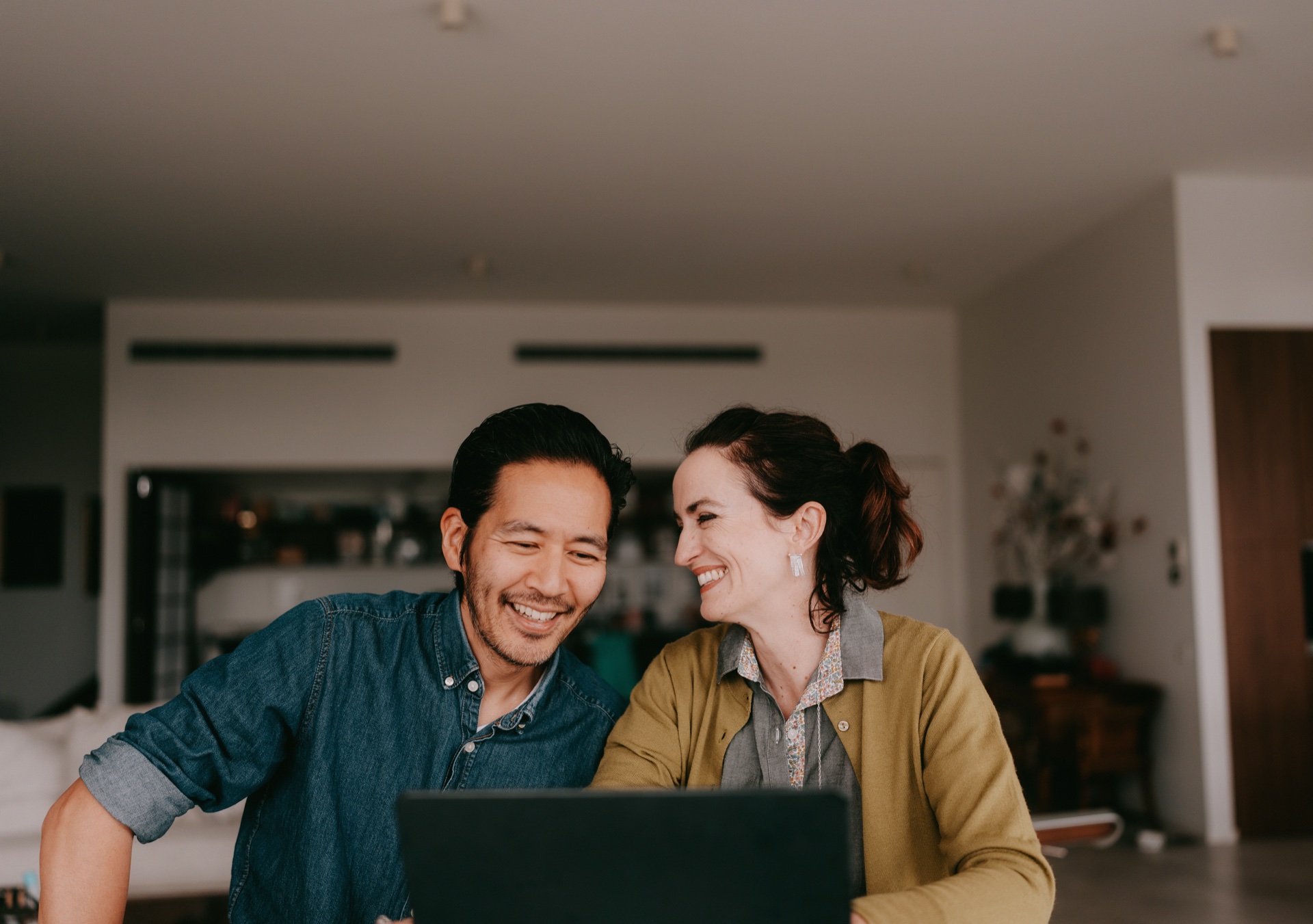 A couple sits together at home looking at a laptop screen and sharing a moment. Online couples therapy through BetterHelp makes it easy to strengthen your relationship from home.