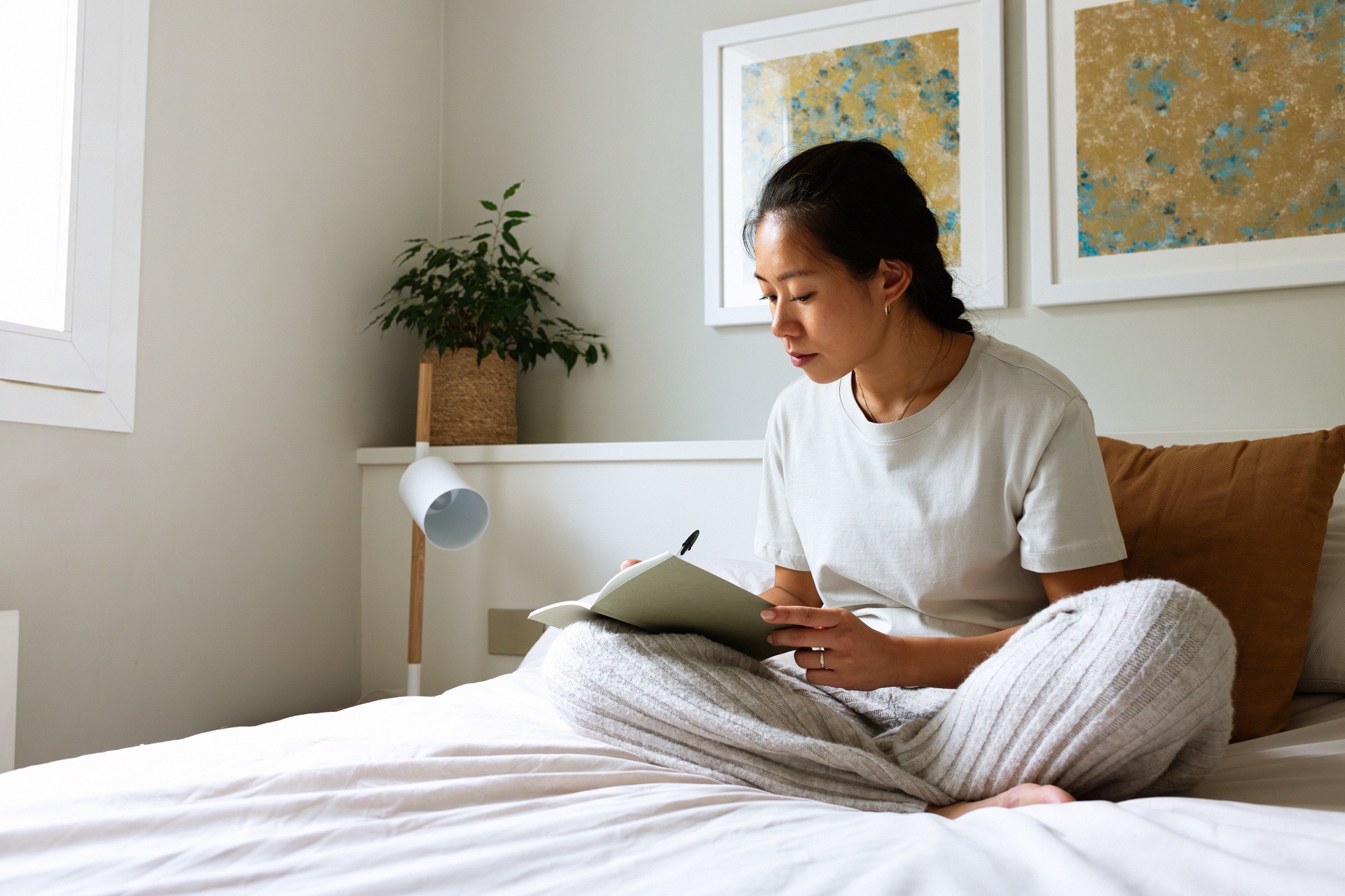 A woman writes thoughtfully in her journal while sitting on her bed. Journaling is a powerful mental health practice that pairs well with online therapy and counseling through BetterHelp.