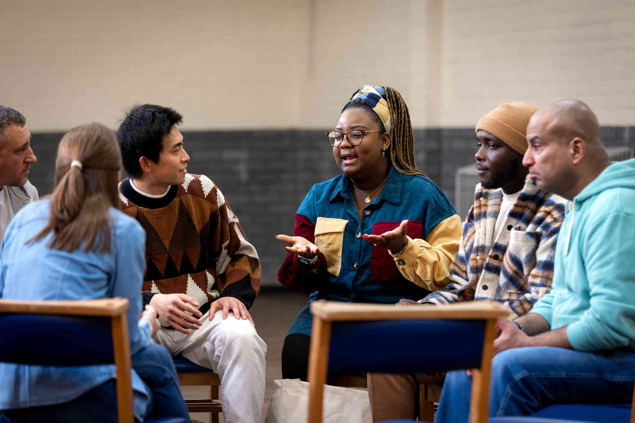 Six adults sit in chairs in a circle during a group therapy session as one woman talks.