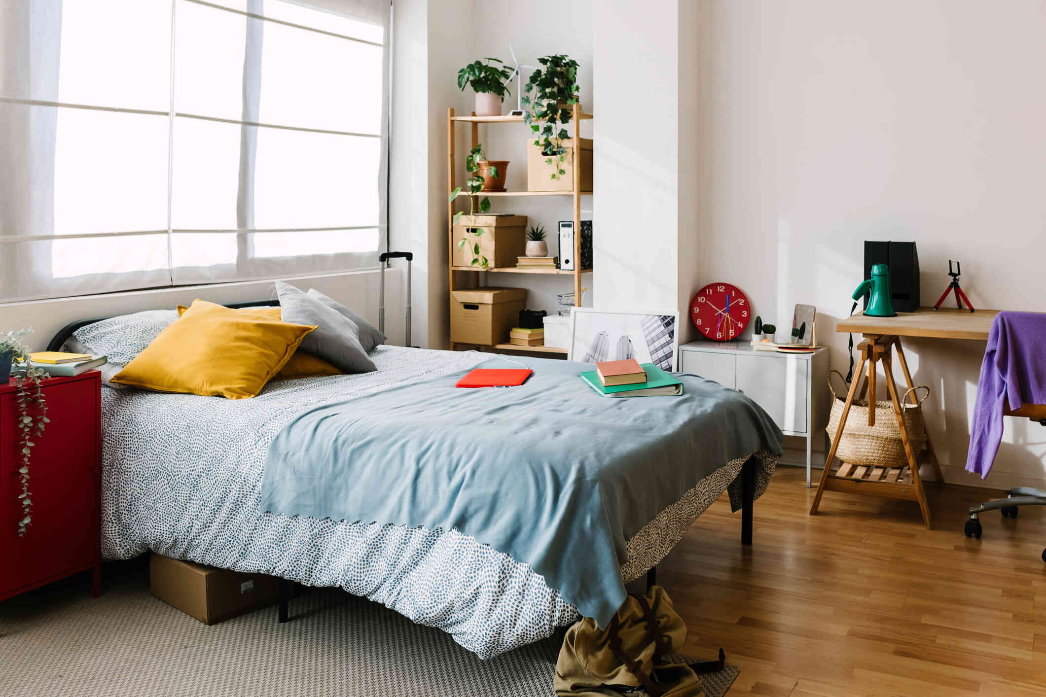 A disorganized bedroom that contains a computer on the bed and clothes on the floor