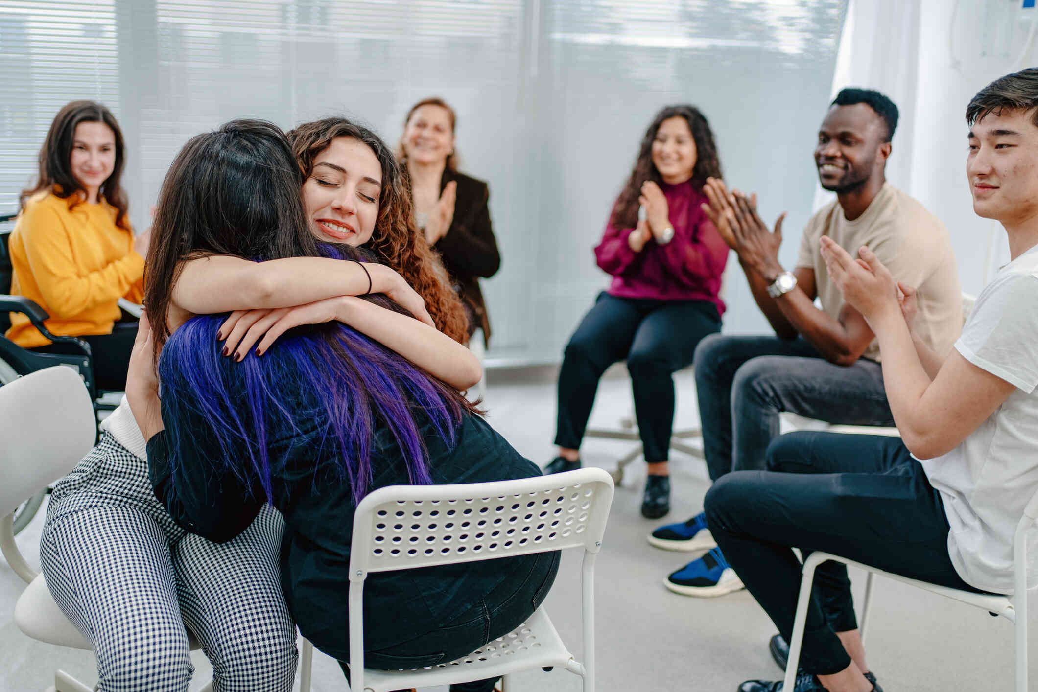 Two woman shug while sitting in a therapy curcle with other adults as they clap.