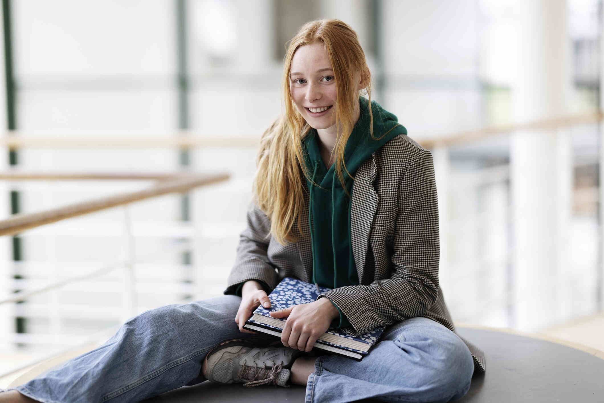 A teen girl in a jackat sits on a table in her school while holding her schoolbooks and smiling at the camera.