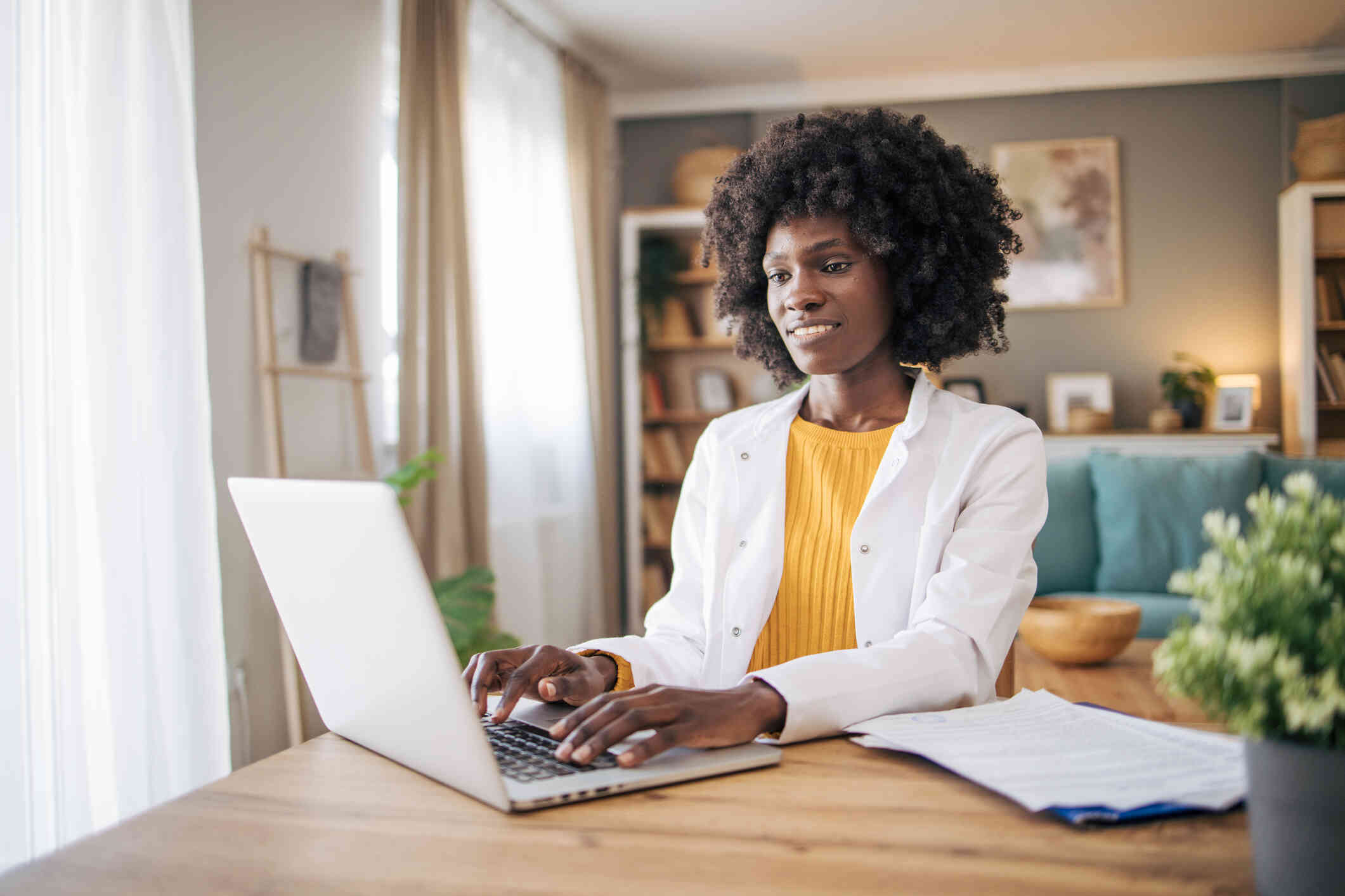 A woman in a yellow shirt sits at a table in her home and types on the laptop open on the table infront of her.