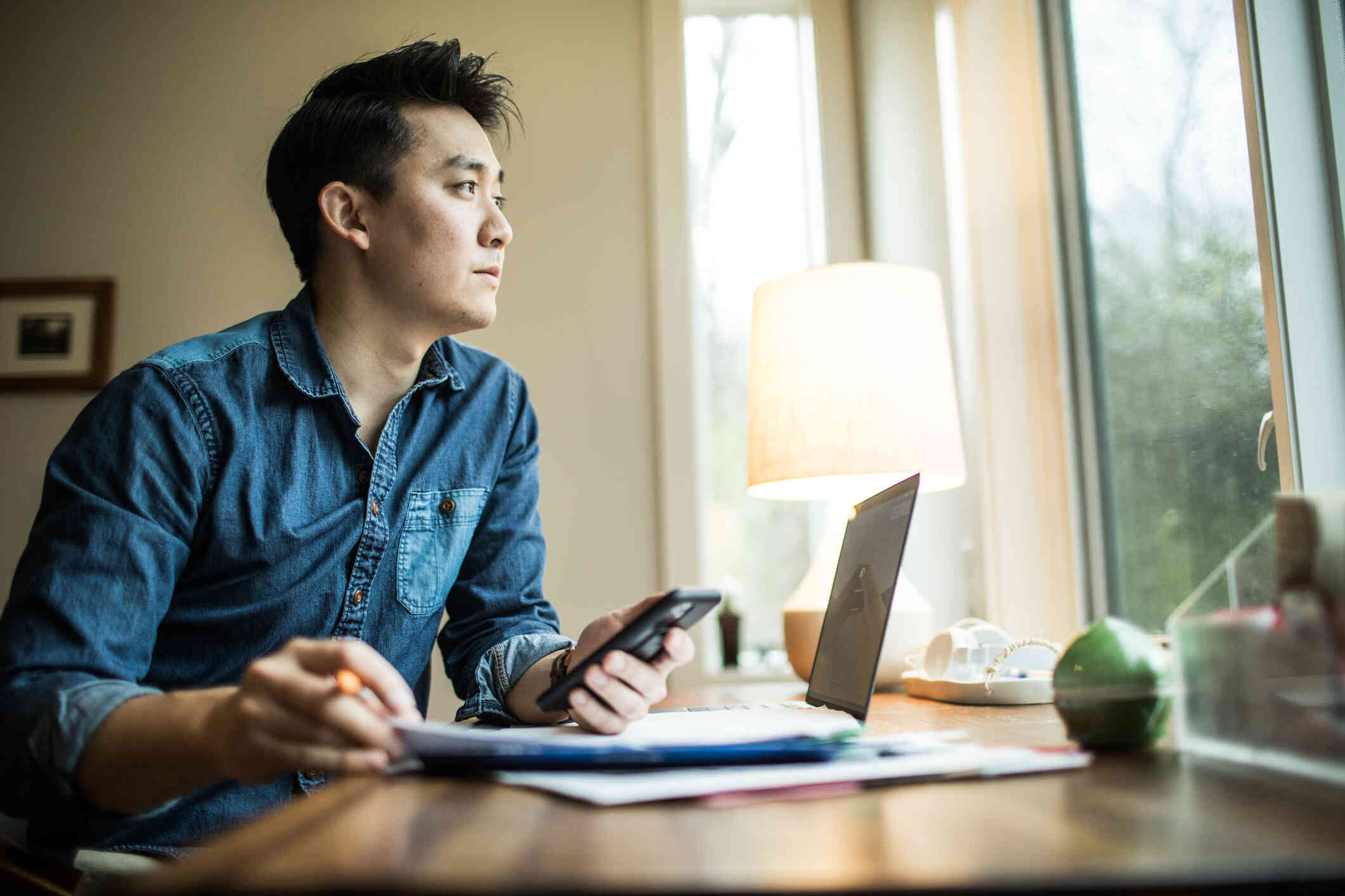A man in a bluejean shirt sits at a table with his laptop and some papers while holding his phone in his hand as he looks out of the window infront of with with a serious expression.
