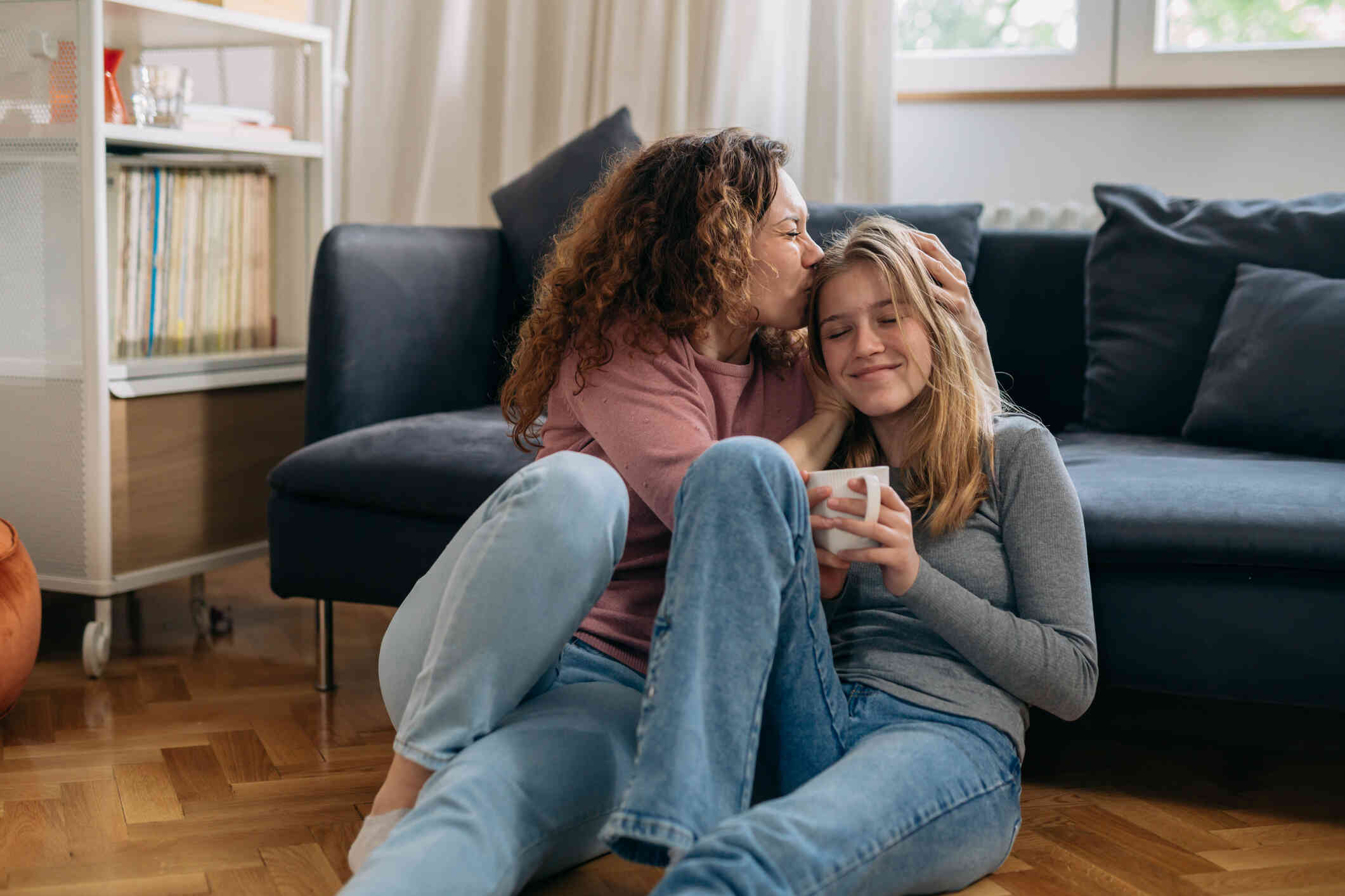 A mom with curly hair kisses the head of a teen girl with blonde hair who is smiling while holding a coffee mug.