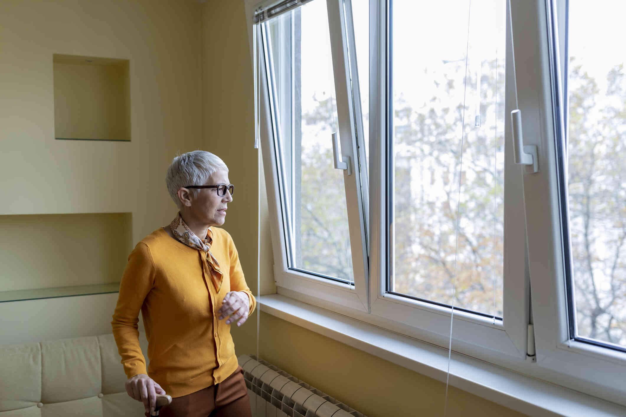 A mature woman in an ornage shirt sitands near a winder in her home and gazes out while deep in thought.