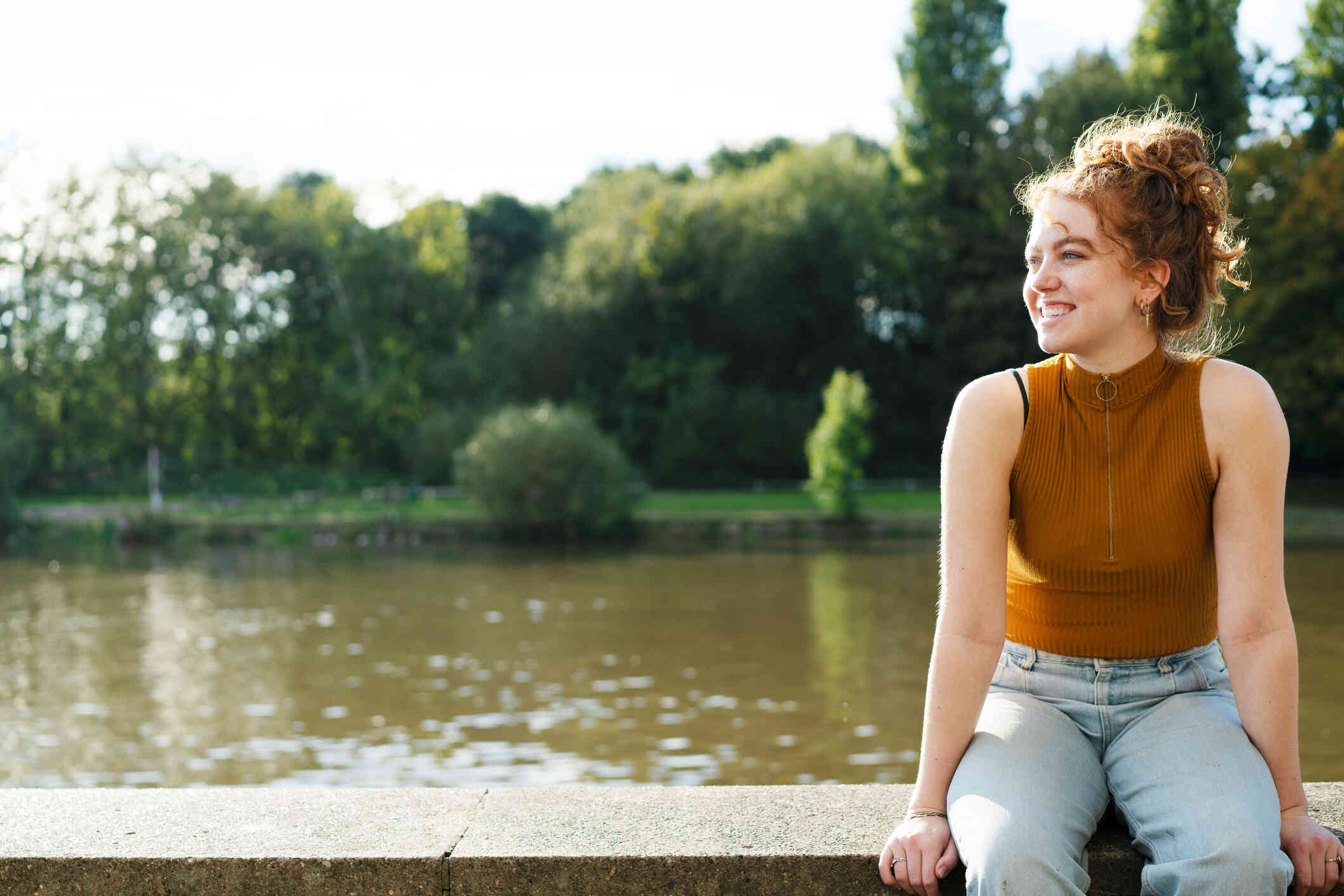 A woman in an orange top sits on a cement ledge outside near a river and smiles while gazing off.