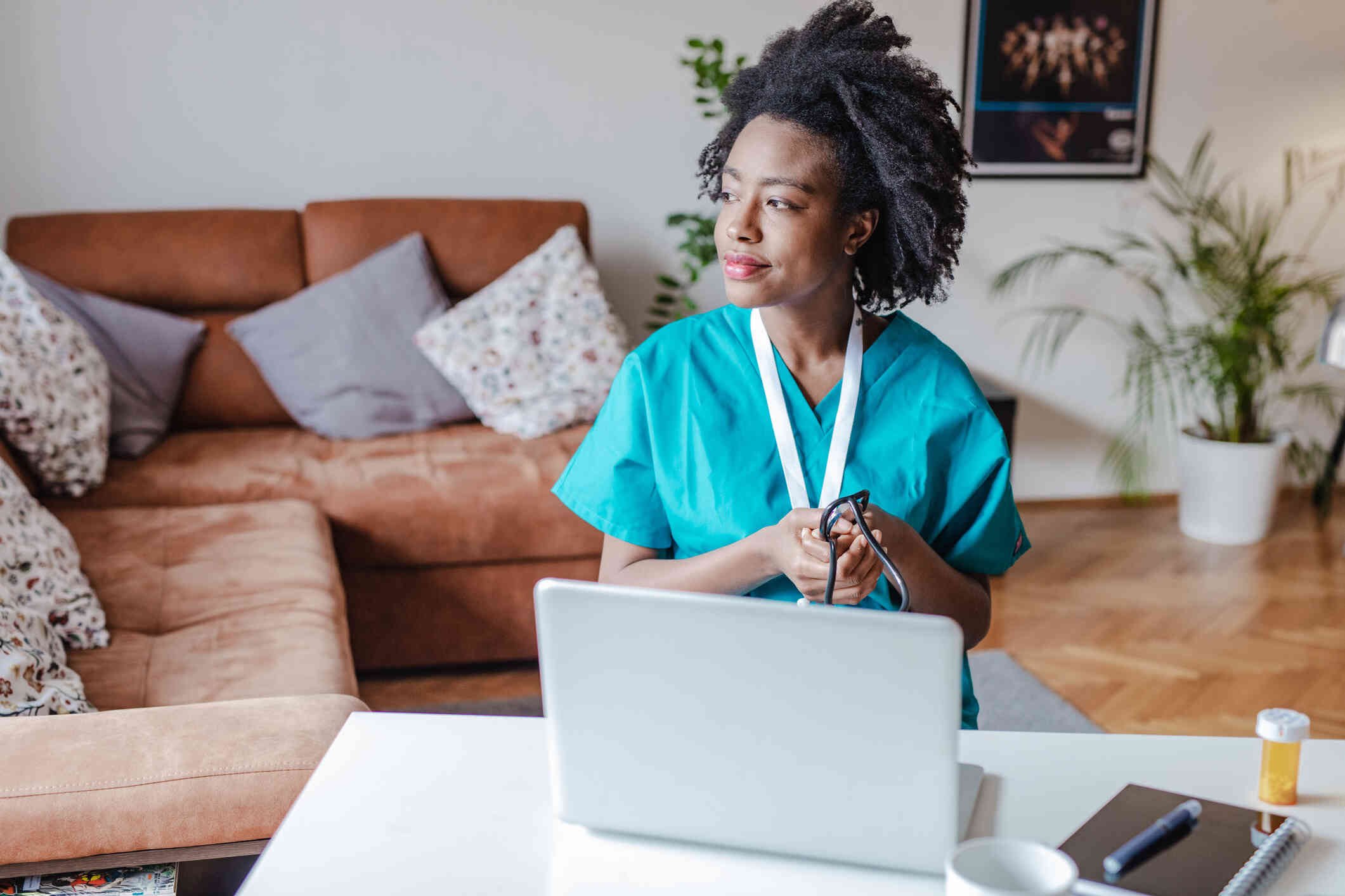 A woman in blue scrubs sits at a table in her home with her laptop open on the talbe infront of her as she gazes off deep in thought.