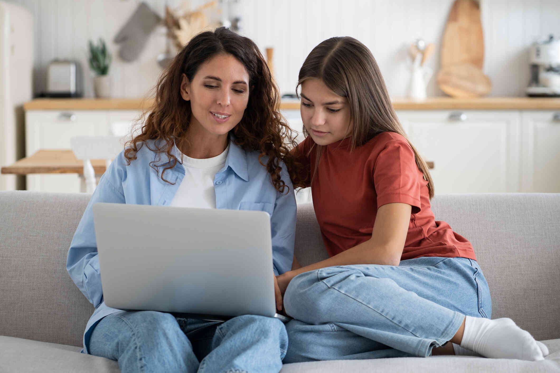 A mother and her daughter sits on a couch while checking something on the laptop.