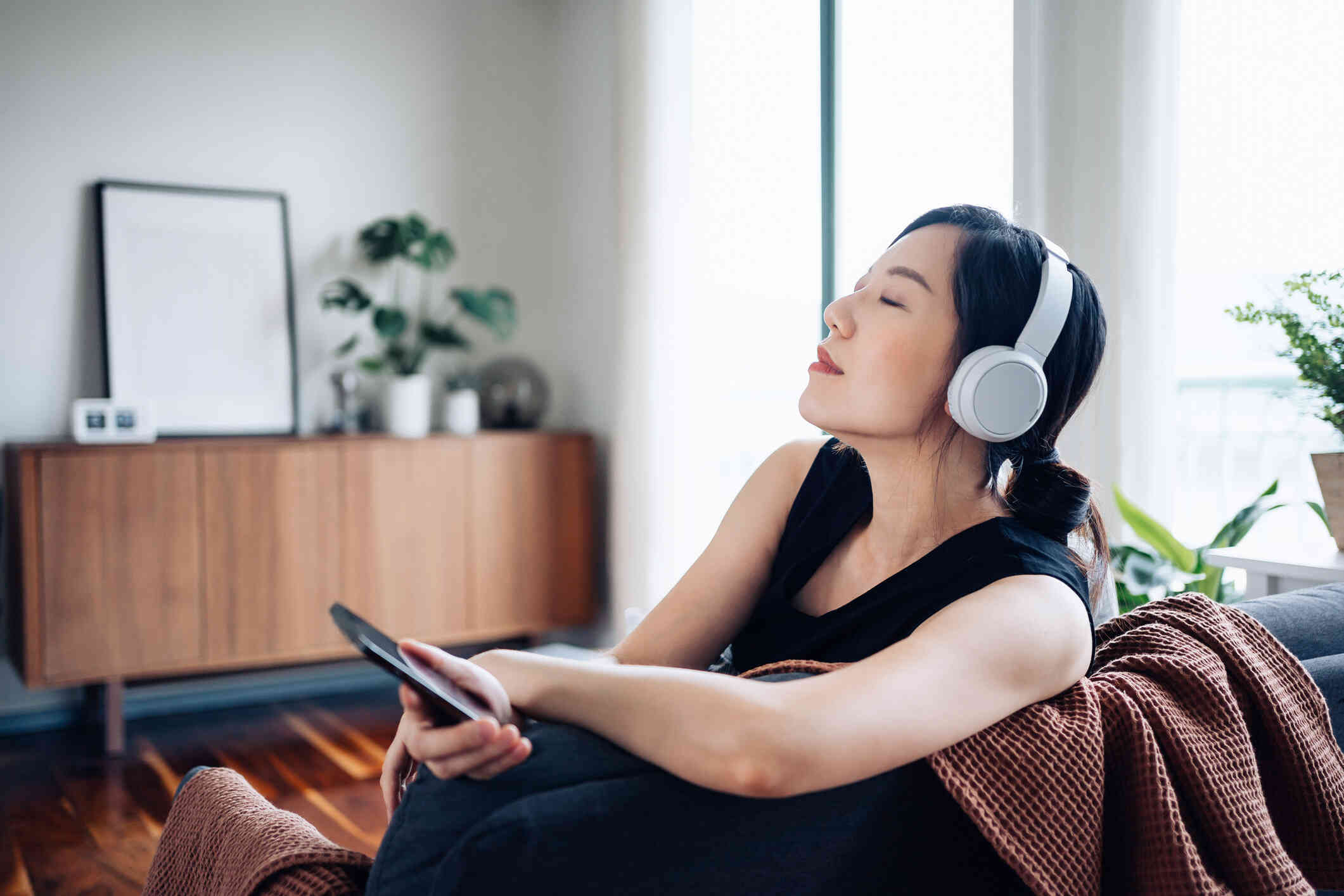 A woman in ablack shirt sits on her couch and closes her eyes while wearing white headphones and holding her phone in her hand.