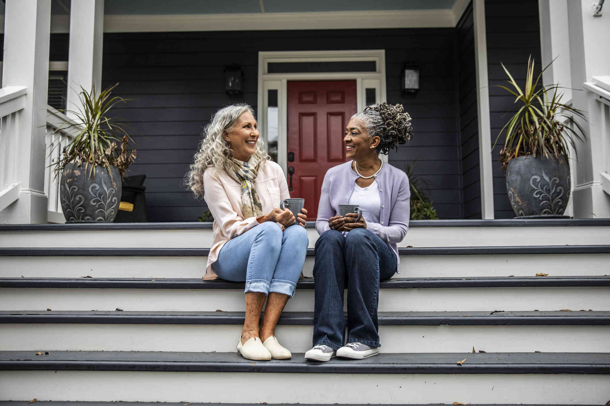 Two mature woman sit on the front porch steps and smile at one another while talking.