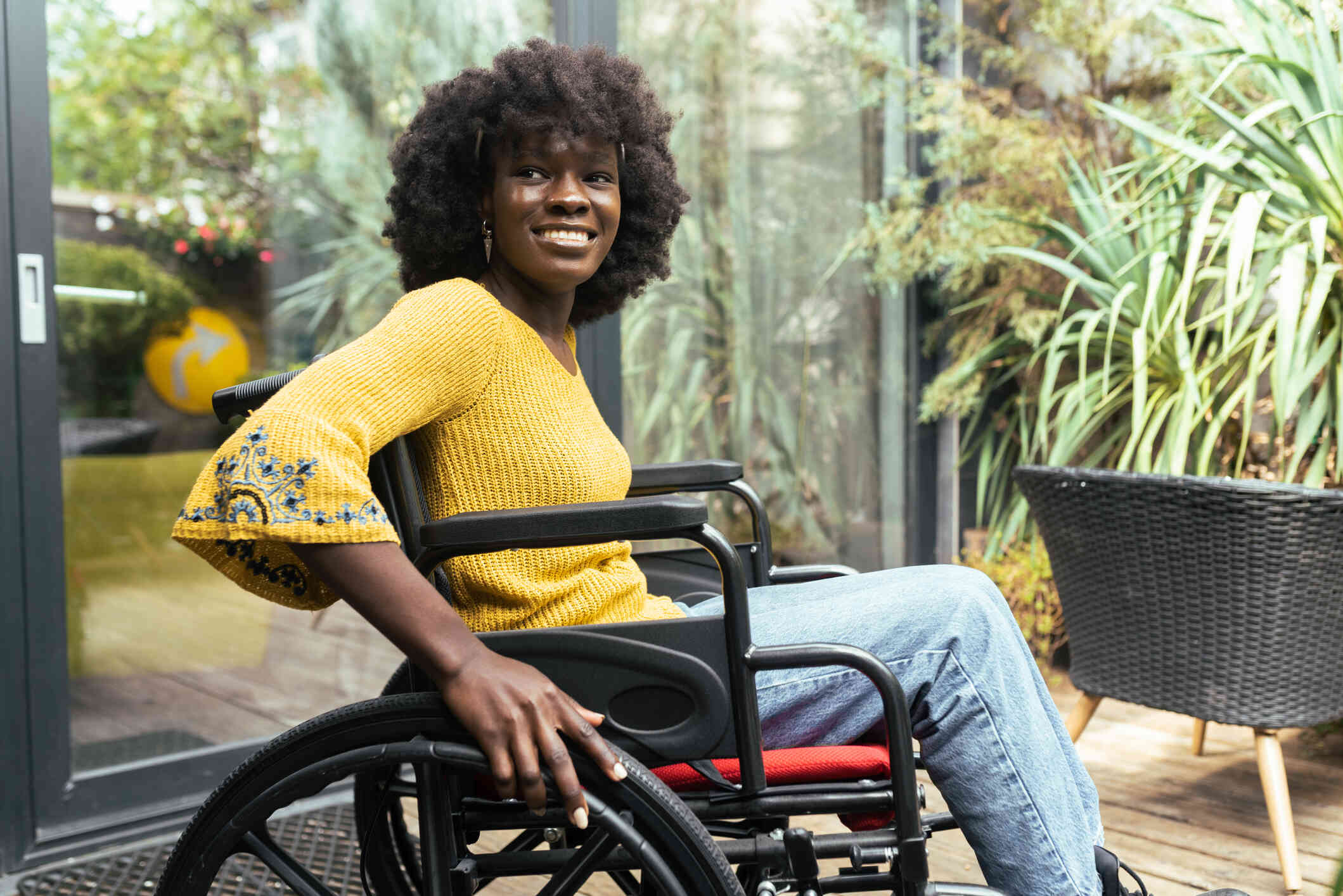 A woman in a yellow shirt sits in a wheelchair outside on a sunny day and smiles while gazing off.