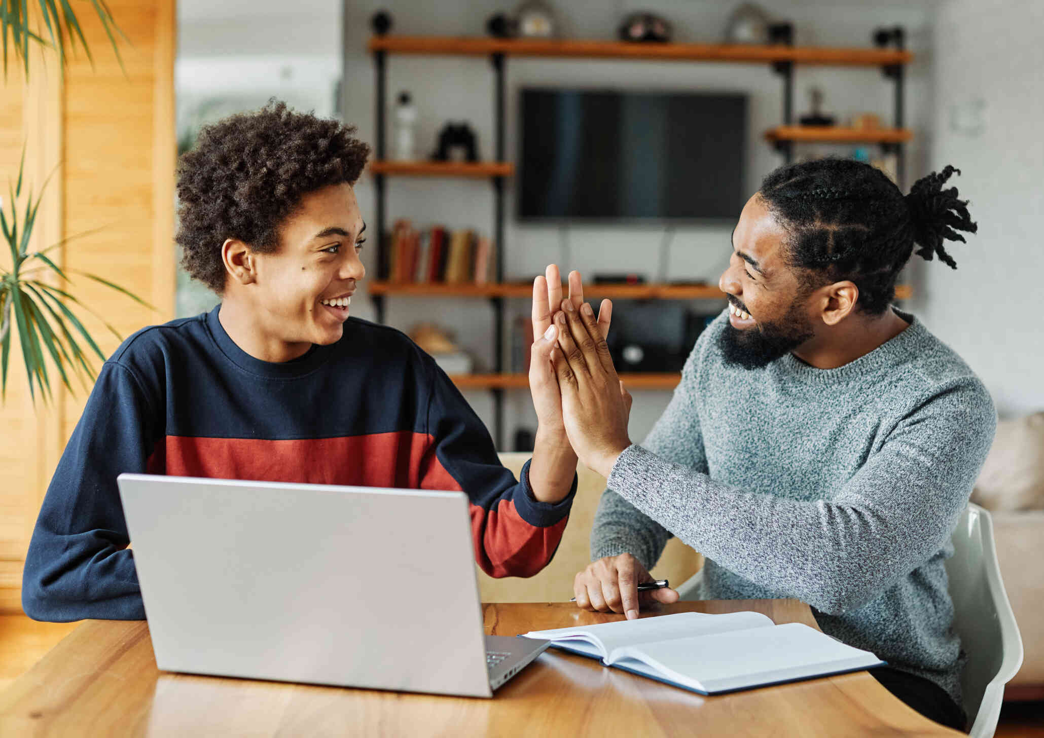 A teenage boy in a red and blue sweatshirt smiles as he high fives an older man with a beard sitting next to him. They are sitting at a living room table with a laptop and notebook in front of them.